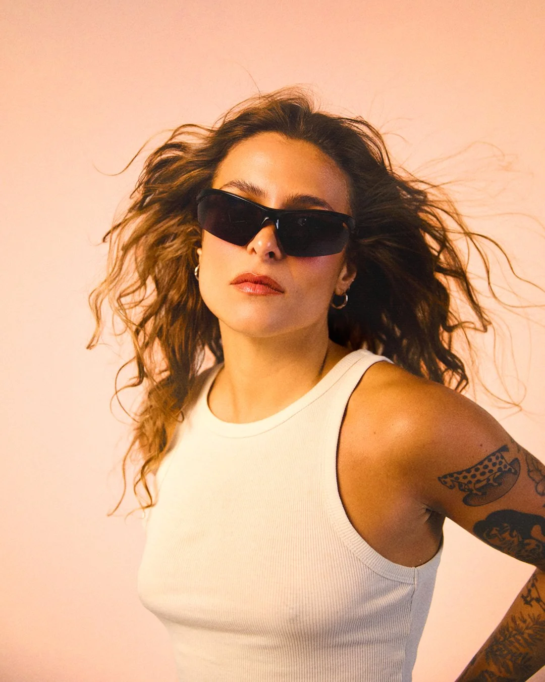 Mulher com cabelos cacheados e óculos escuros, vestindo regata branca, com tatuagens no braço, posando contra fundo claro.
Woman with curly hair and sunglasses, wearing a white tank top, with tattoos on her arm, posing against a light background.