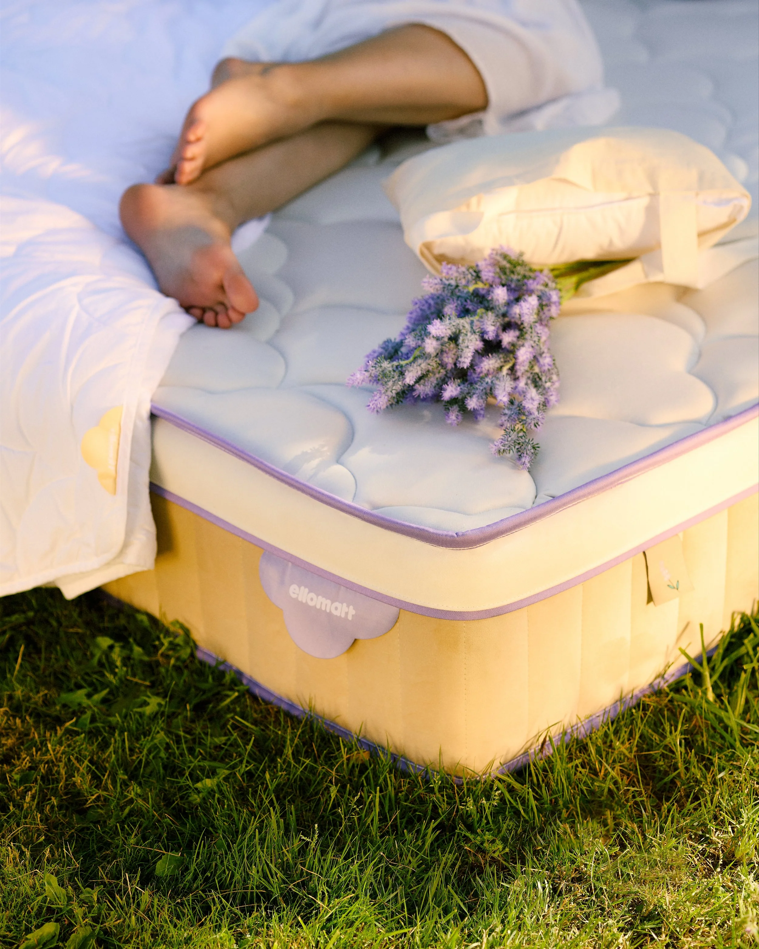 A mattress on grass with a pillow, a bouquet of purple flowers, and a person sleeping on the side with bare legs visible.