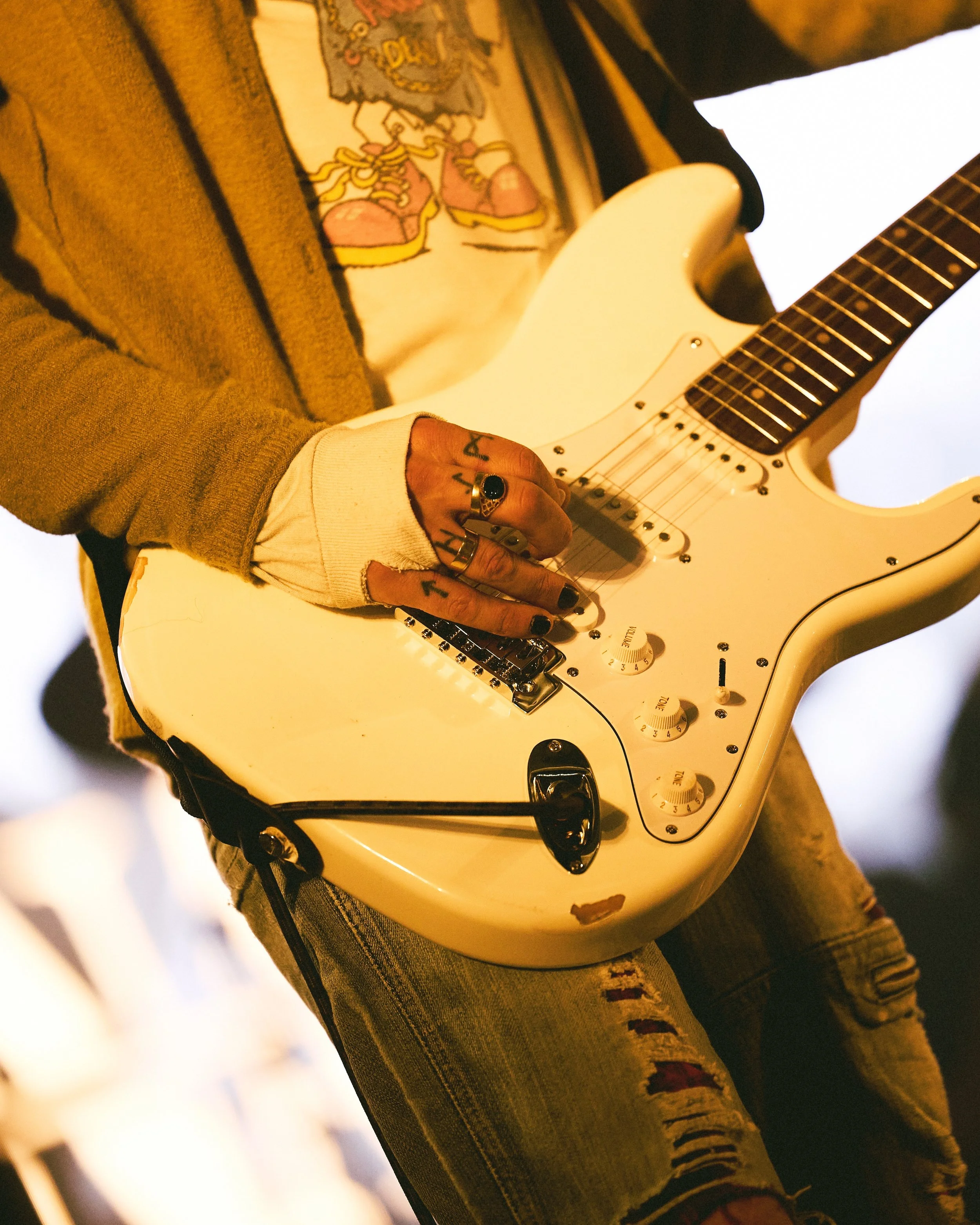Person holding a white electric guitar, wearing a brown coat and a graphic t-shirt, as well as ripped jeans, with visible tattoos on the hand.