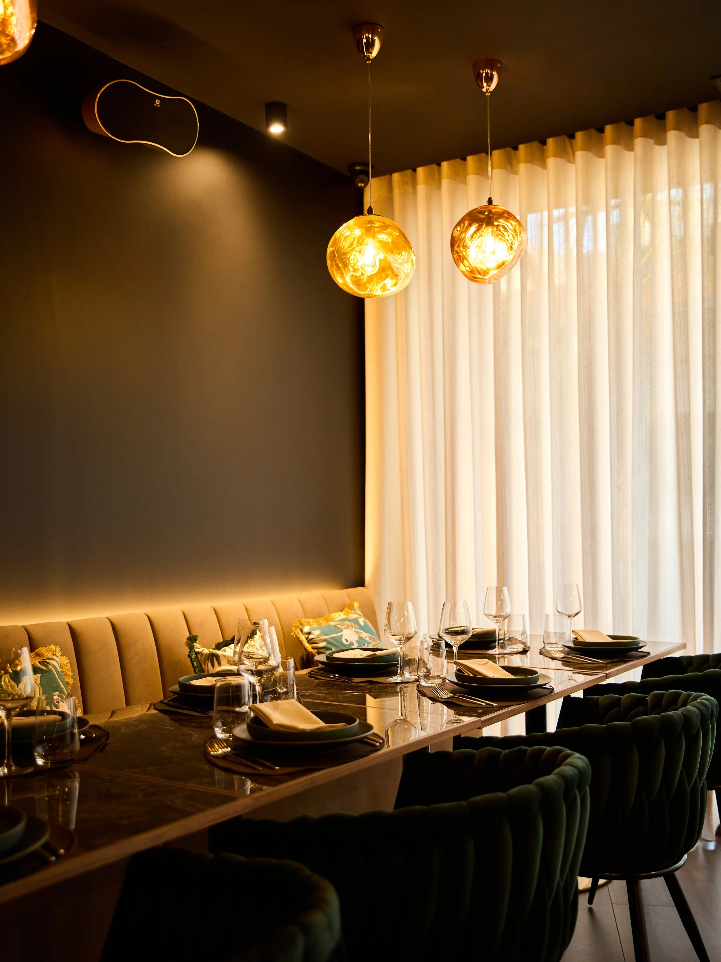A modern dining room with a long table set for a meal, featuring black plates, wine glasses, and cloth napkins. The room has dark walls, a beige cushioned bench along the wall, velvet green chairs, sheer curtains, and two hanging pendant lights with golden glass shades.