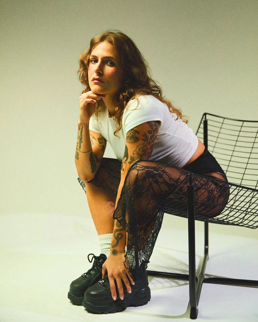 Mulher com tatuagens, cabelos ondulados, roupa branca, saia de renda preta, sapatos preta, sentada em uma cadeira de arame
Woman with tattoos, wavy hair, white outfit, black lace skirt, black shoes, sitting on a wire chair