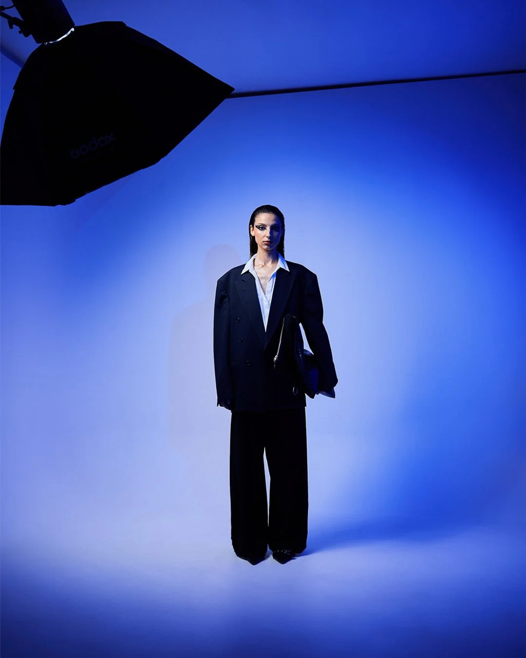 Mulher vestida com terno preto e camisa branca, de pé em fundo azul, iluminada por luz de estúdio.
Woman dressed in black suit and white shirt, standing on blue background, illuminated by studio light.