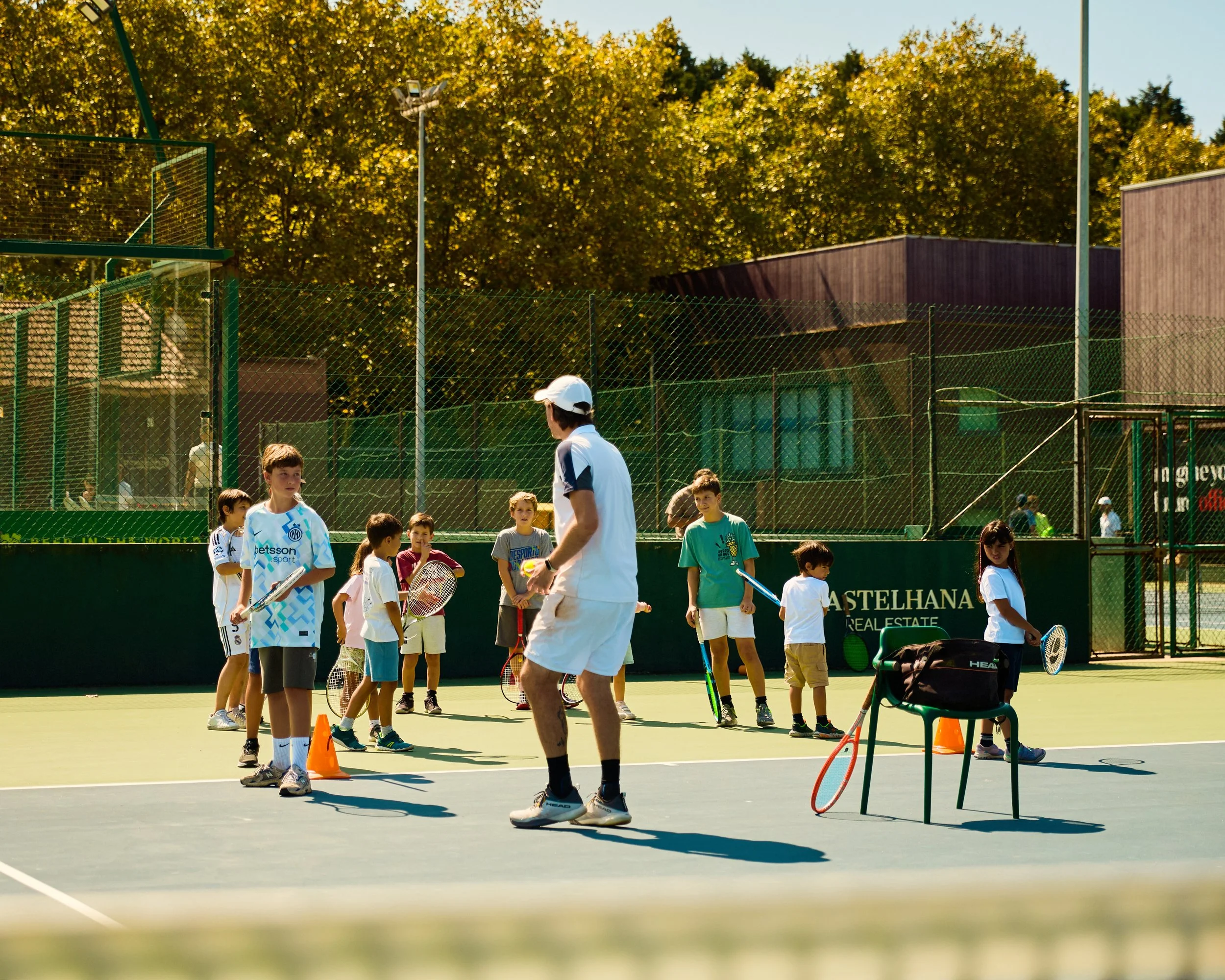 Children participating in a tennis lesson on an outdoor court. An instructor is demonstrating, with children holding tennis rackets, and orange cones marking areas. Background includes a chain-link fence, trees, and a building.