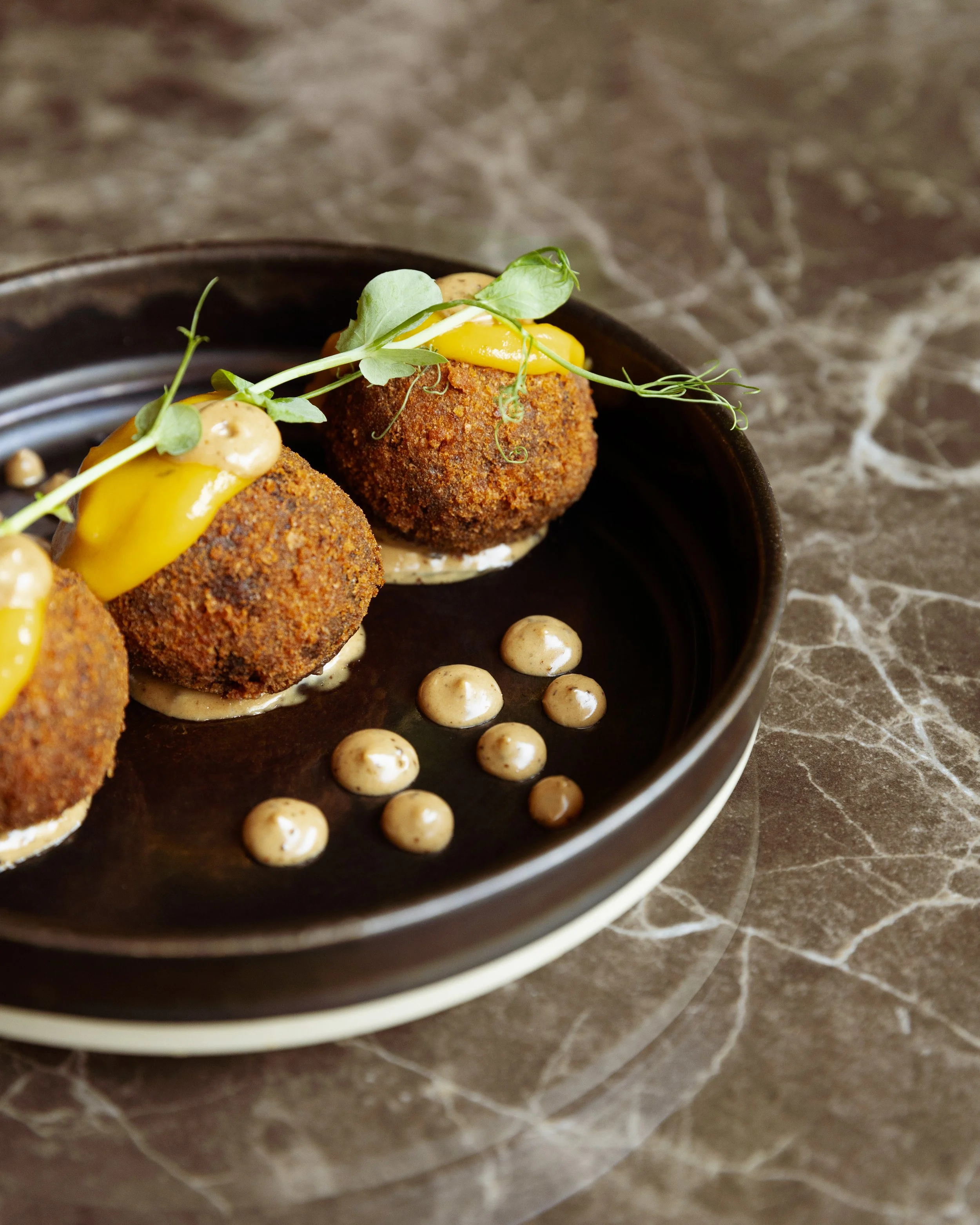 Three breaded and fried balls topped with a yellow sauce and microgreens, served on a black plate with dotted creamy sauce around them.