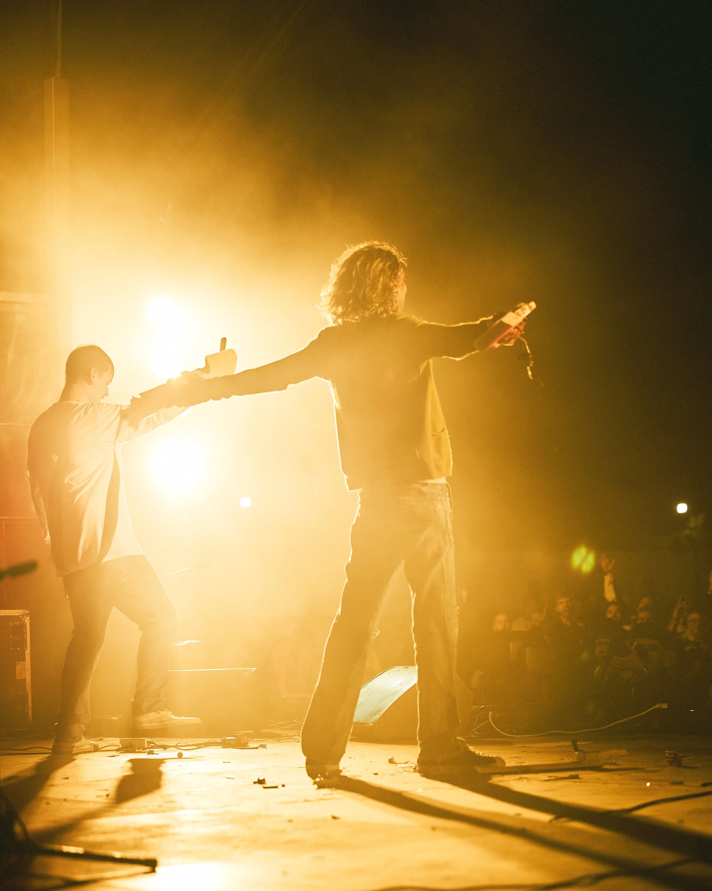 Two musicians on stage performing at night, one with long curly hair holding a microphone, the other with short hair holding a smartphone, both illuminated by bright yellow stage lights.