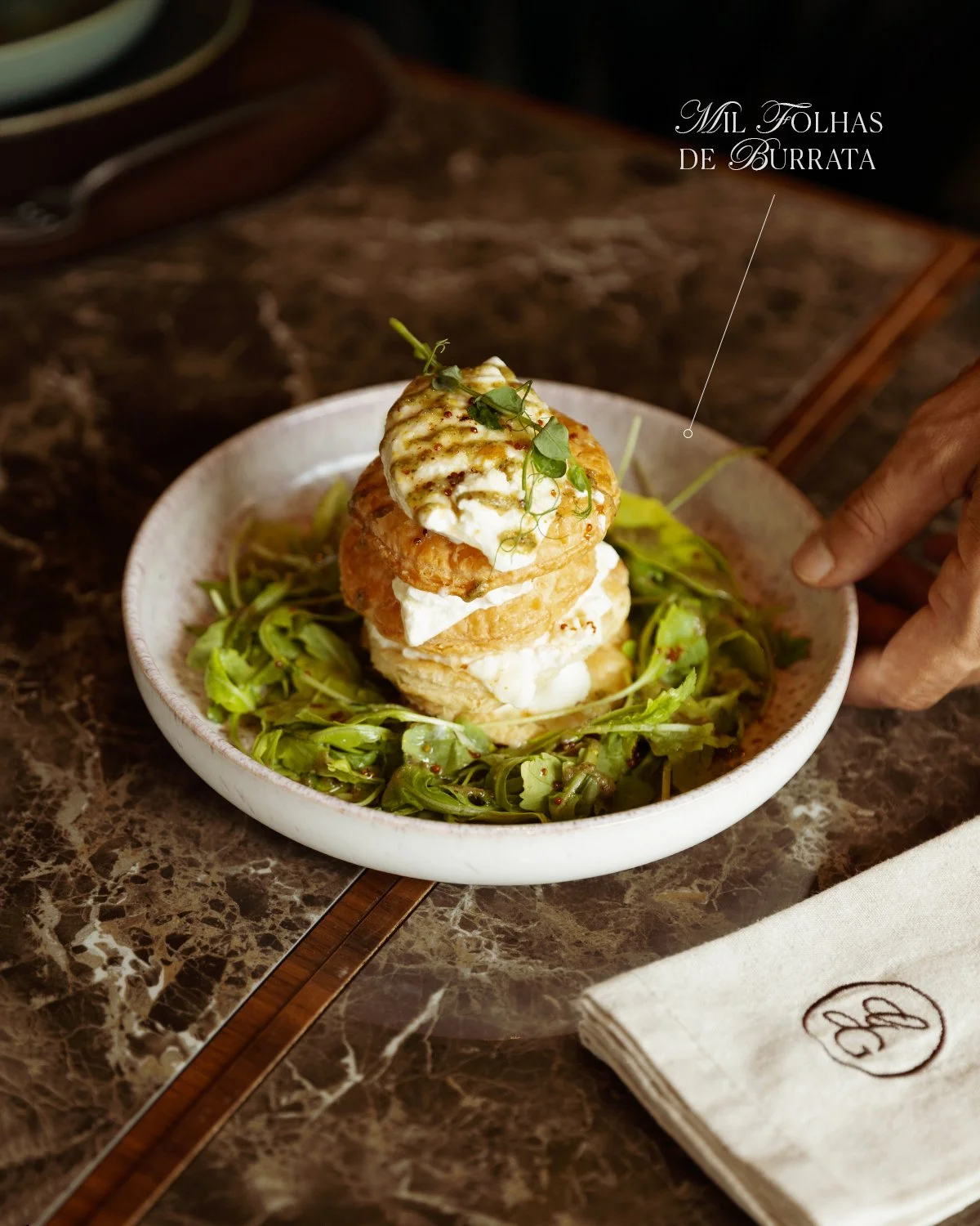 A dish called 'Milk Folhas de Burrata' featuring layered fried bread with burrata cheese and garnished with microgreens, served on a bed of arugula in a white bowl on a dark marble table.