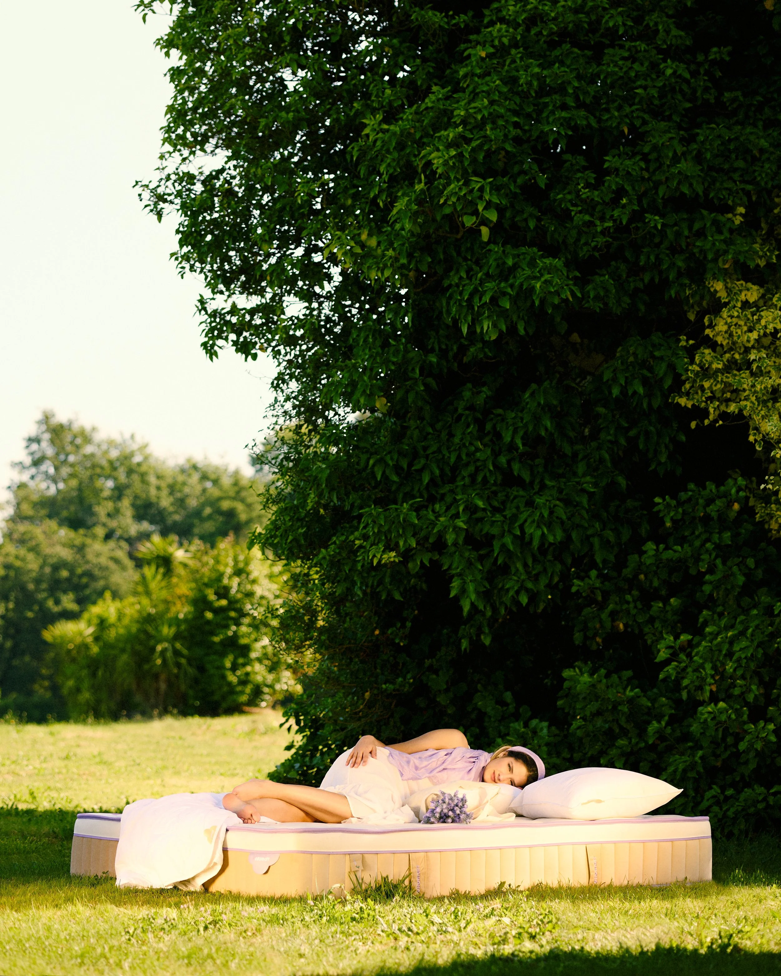 A woman wearing a lavender dress and white headband, lying on a mattress outdoors under a large bush, resting her head on a pillow with a bouquet of purple flowers nearby.