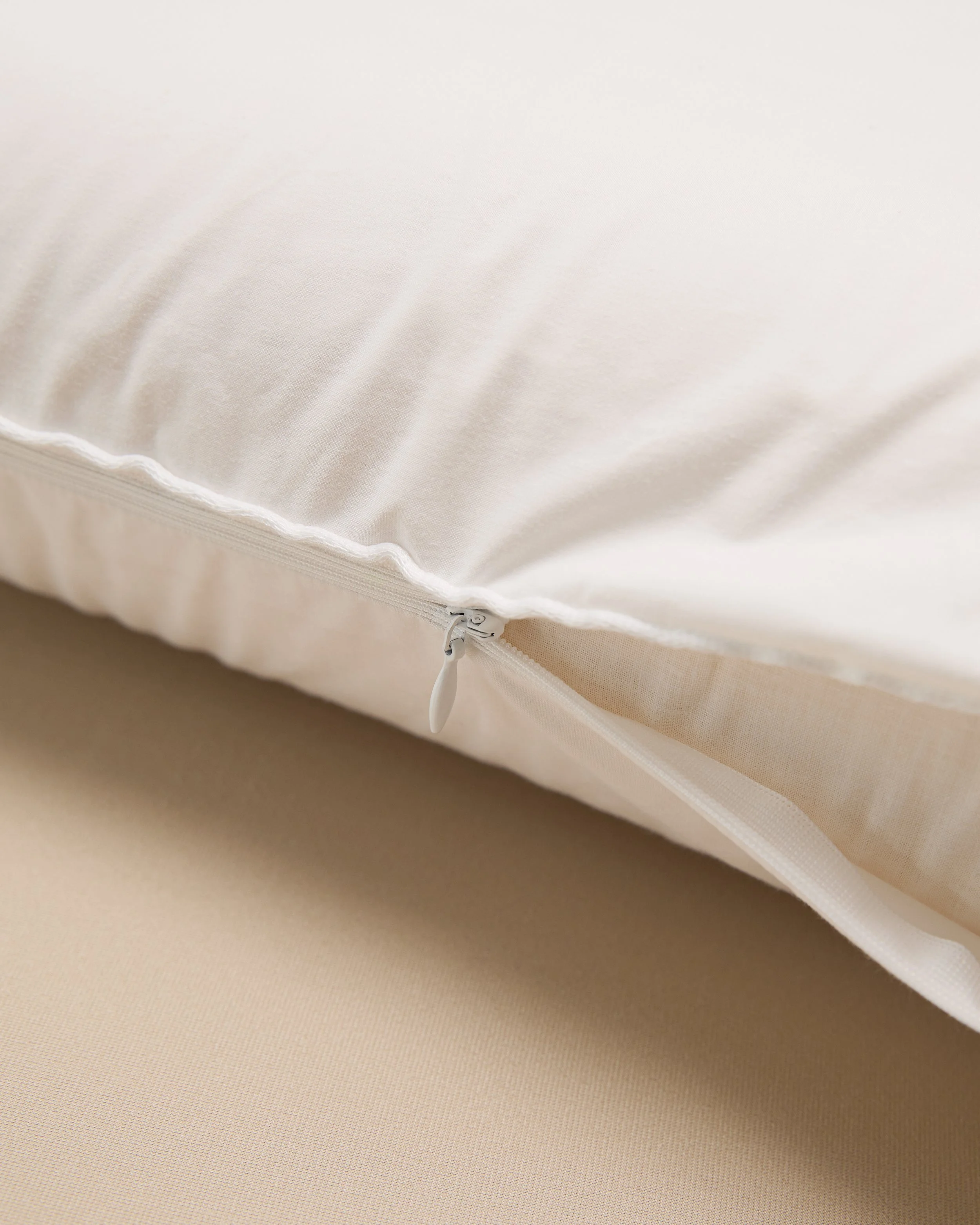 Close-up of a white pillow with a zipper, lying on a beige surface.
