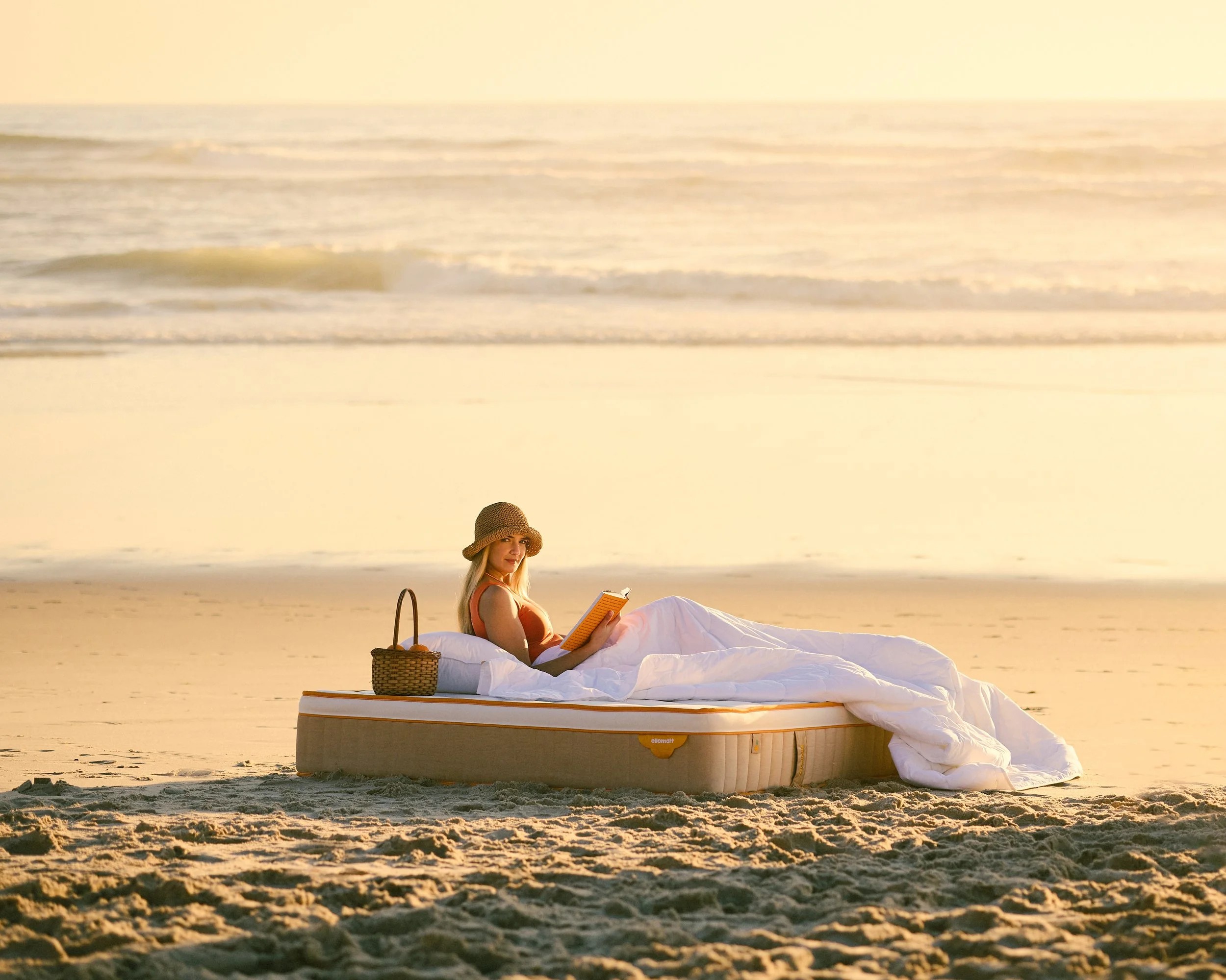 A woman relaxing on an air mattress on the beach, reading a book, with a straw hat and a woven bag, during sunset.