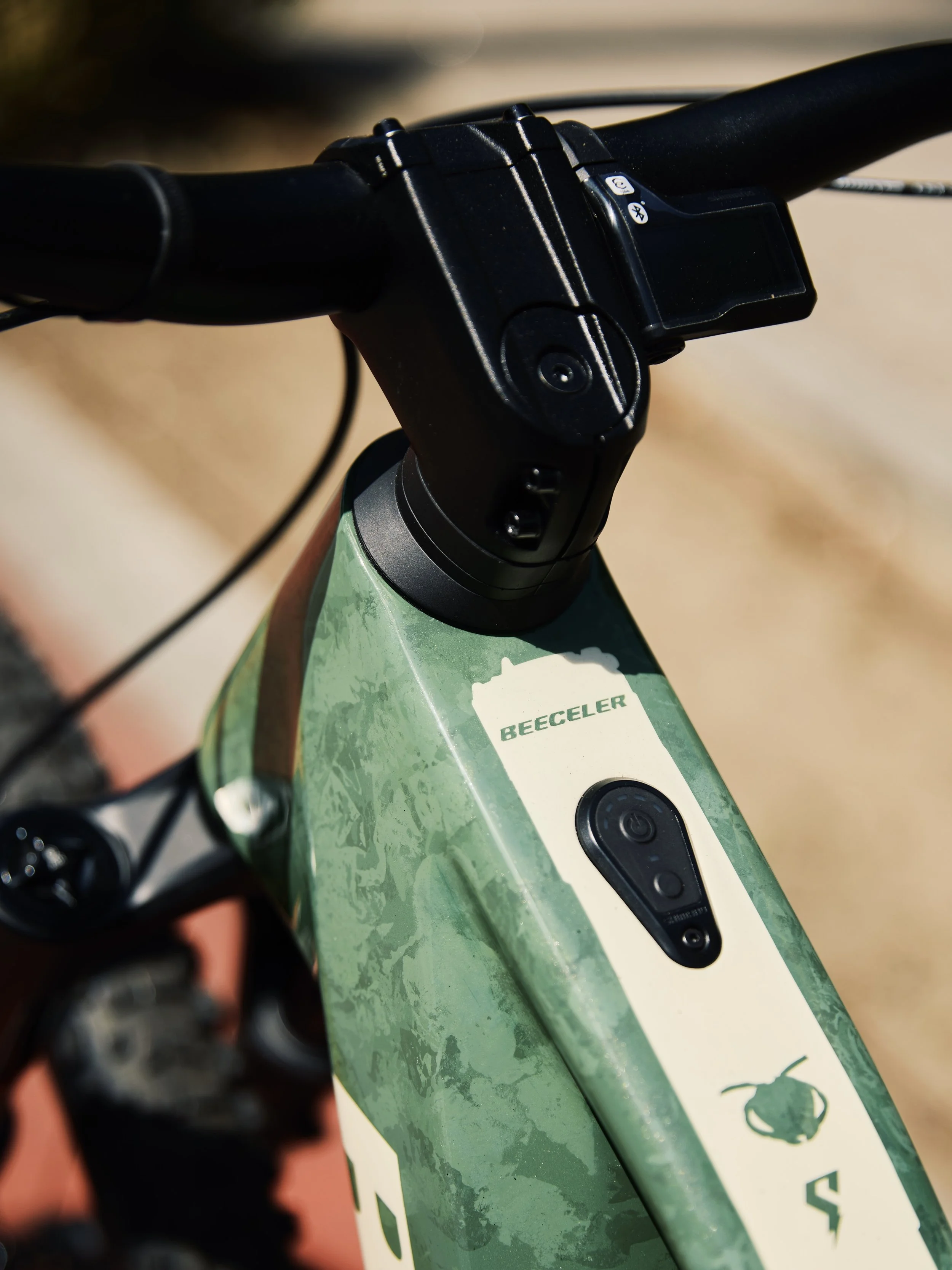 Close-up of an electric bicycle handlebar and top tube showing the brand BeeCeler and a camera on the frame.