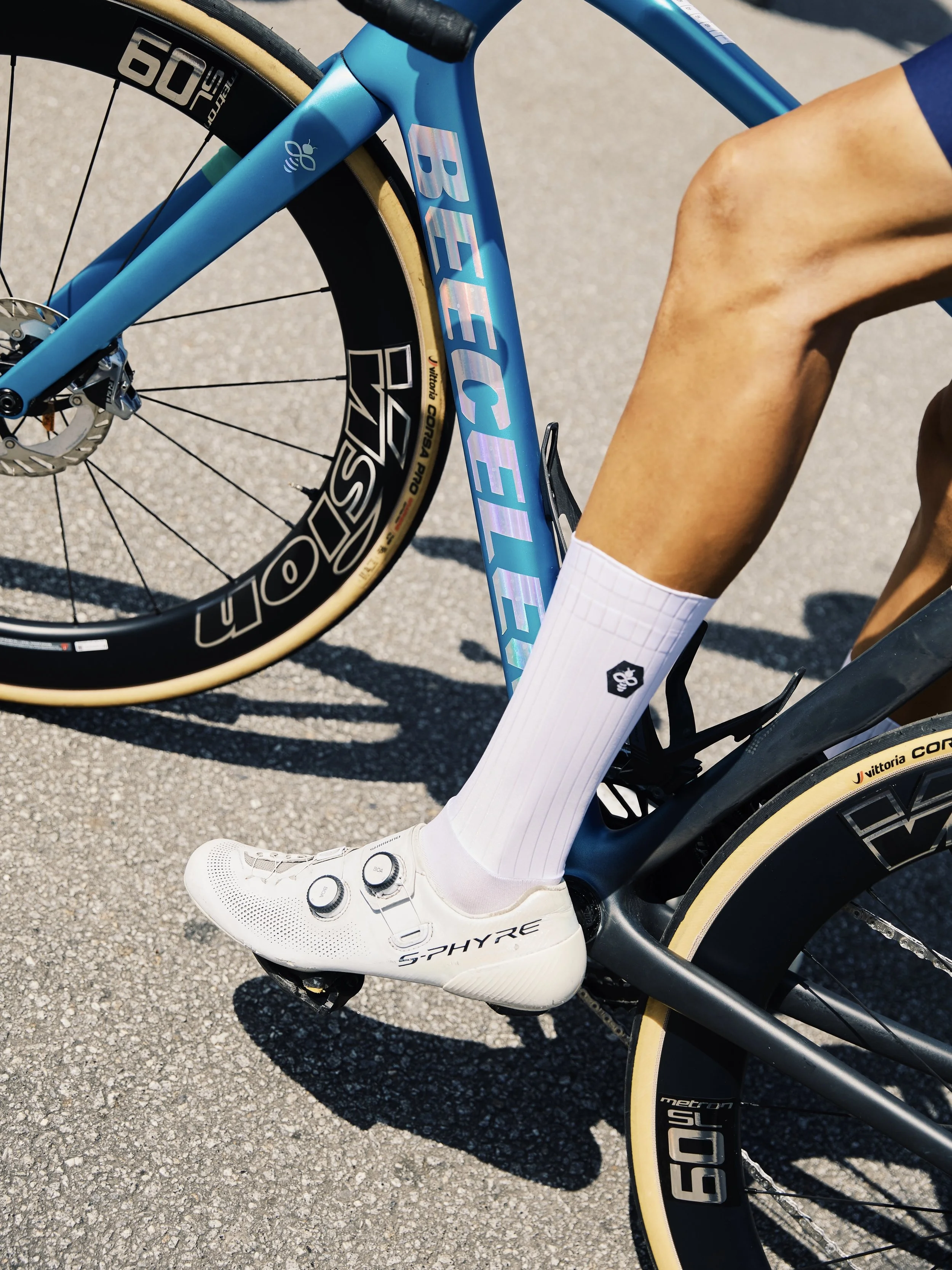Close-up of a cyclist's leg and foot on a blue BeeCome bike, wearing white cycling shoes and white socks, on a paved road.