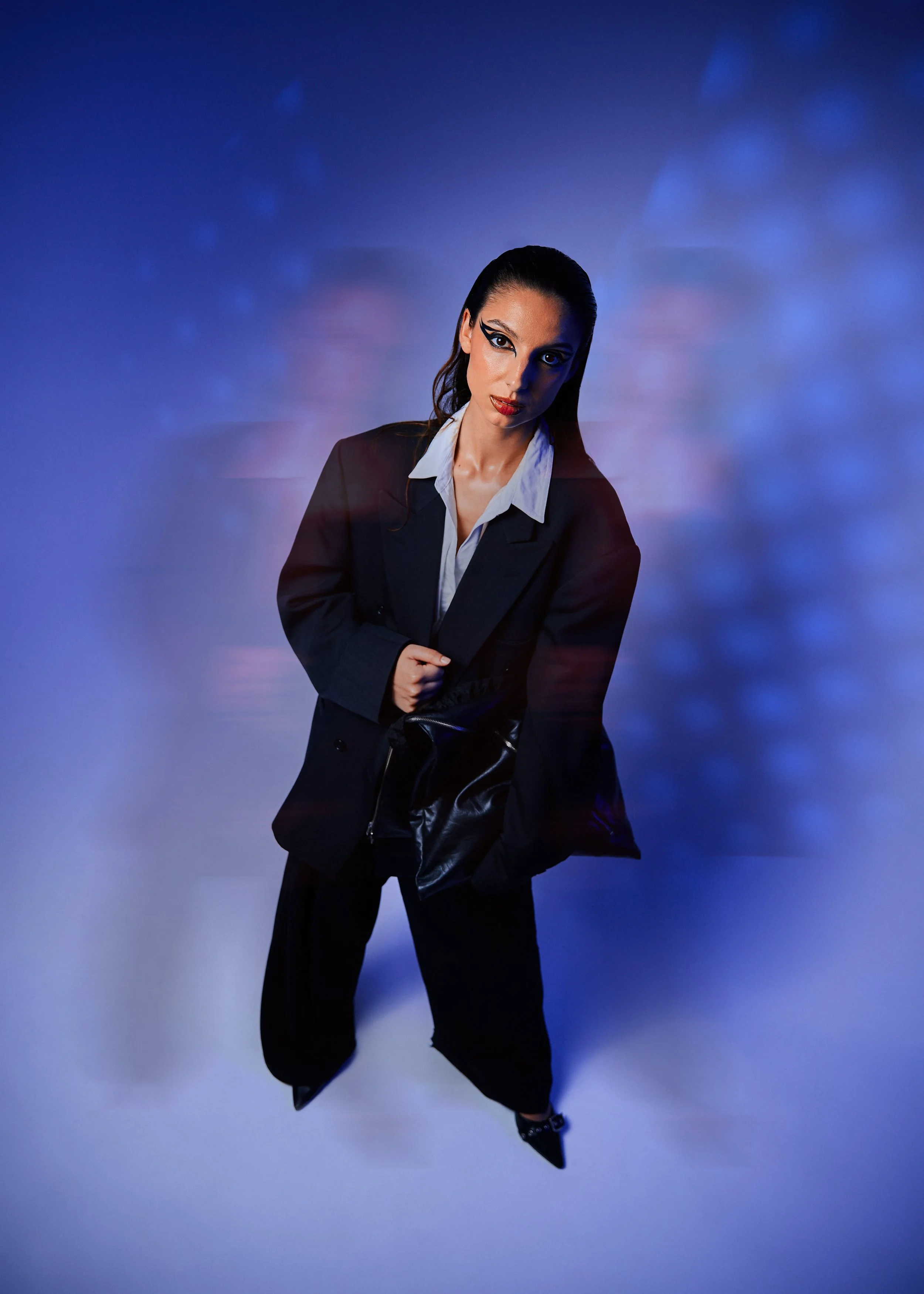 Mulher com maquiagem marcante, cabelo preso, vestindo terno preto e camisa branca, segurando uma bolsa preta, contra fundo azul com efeito de movimento.
Woman with striking makeup, hair tied up, wearing a black suit and white shirt, holding a black