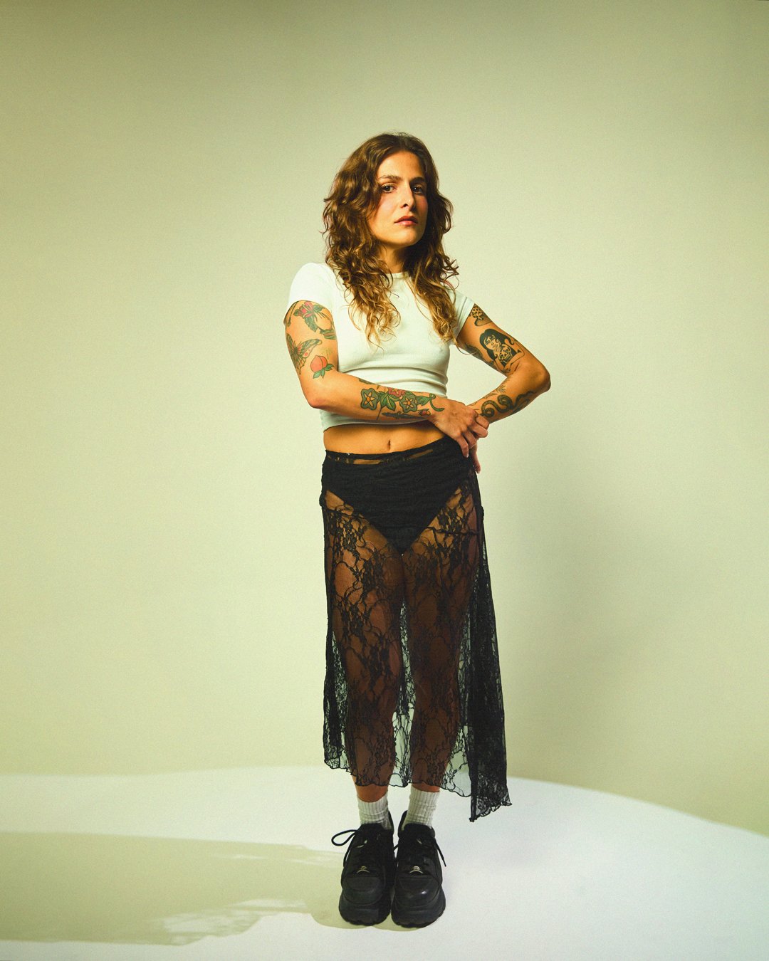 A woman with curly hair and tattoos on her arms standing with her hand on her stomach against a plain background, wearing a white crop top, black lace skirt, and black shoes.