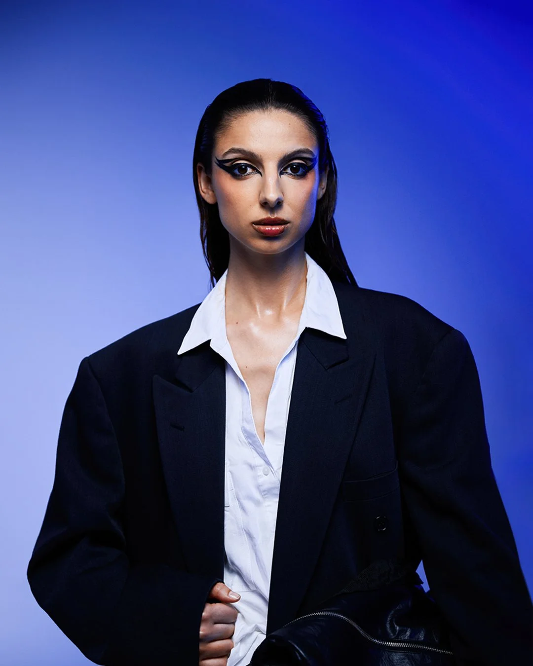 Mulher com maquiagem marcante, cabelo escuro, vestindo terno preto e camisa branca, contra fundo azul
Woman with striking makeup, dark hair, wearing a black suit and white shirt, against a blue background