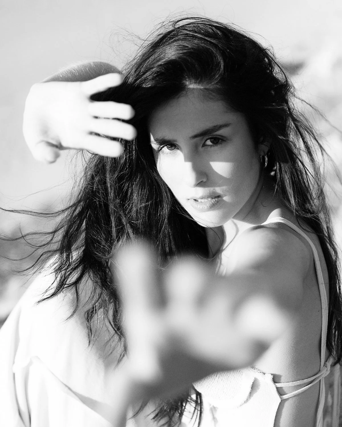 Retrato em preto e branco de uma mulher com cabelo escuro e olhos intensos, olhando para a câmera, com uma mão próximas ao rosto. Ela usa um vestido de alça. Black and white portrait of a woman with dark hair and intense eyes, looking at the camera,