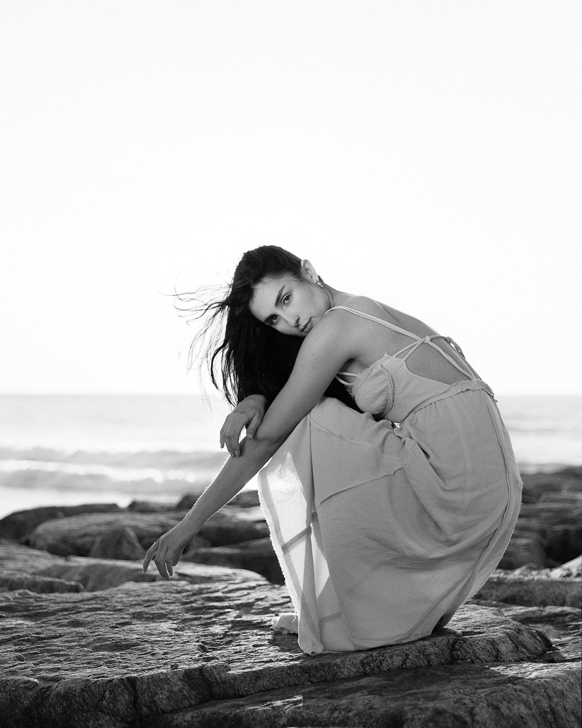 Mulher de cabelo longo e escuro, vestindo um vestido claro, sentada sobre rochas na praia, olhando para a câmera com fundo do mar ao fundo, preto e branco.
Woman with long dark hair, wearing a light dress, sitting on rocks on the beach