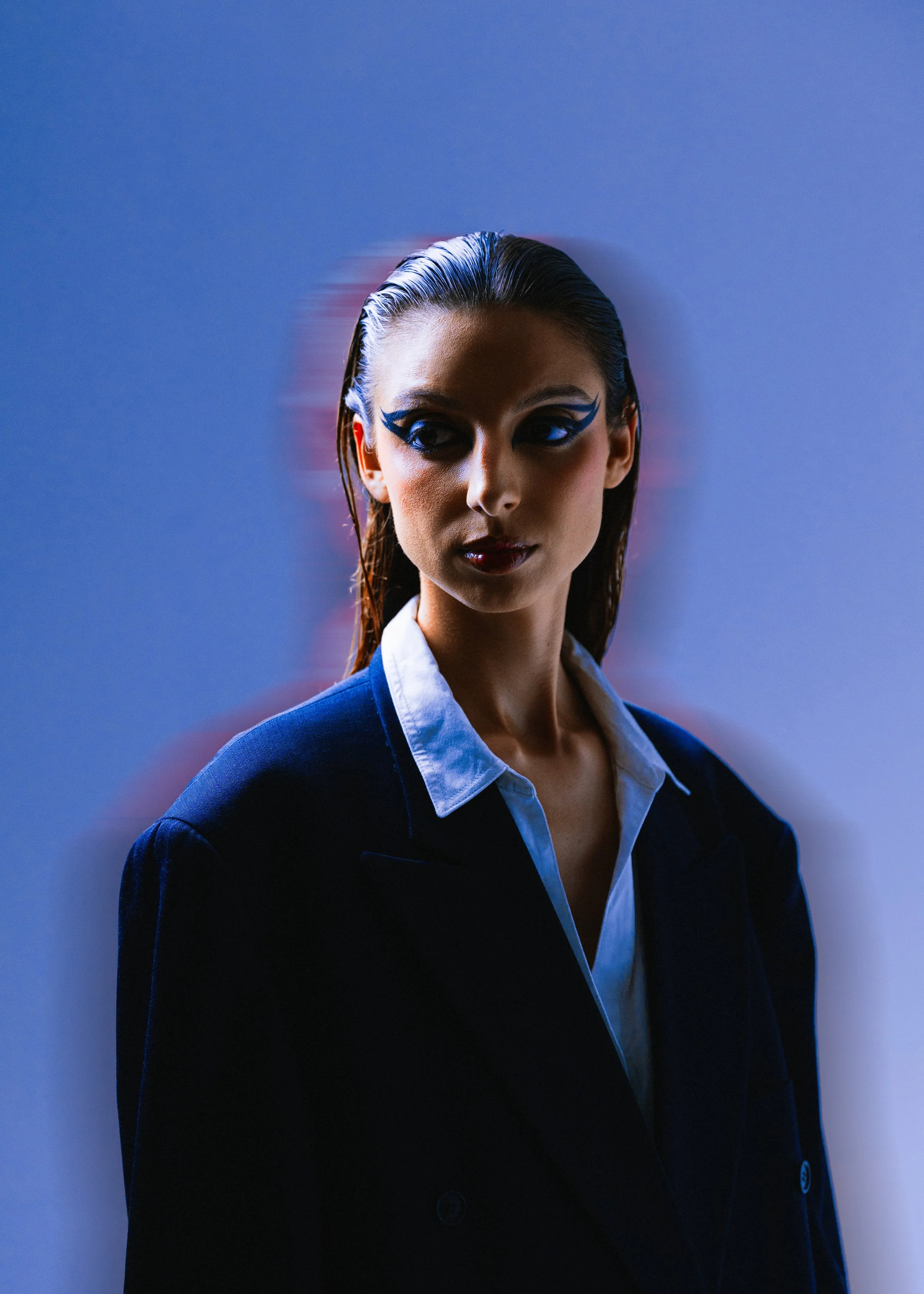 Mulher com maquiagem marcante, cabelo molhado, vestindo jaqueta escura e camisa branca, em fundo azul com efeito de desfoque.
Woman with striking makeup, wet hair, wearing a dark jacket and white shirt, on a blue background with a blur effect.