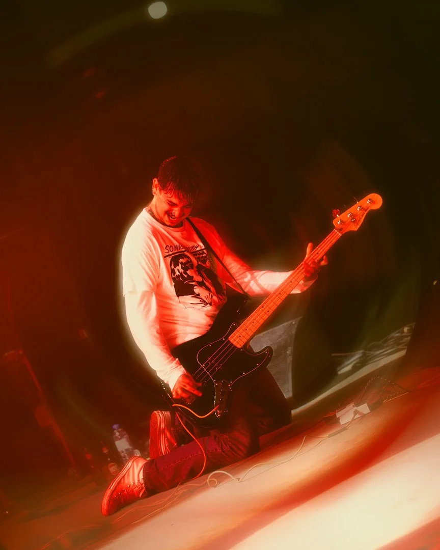 Person kneeling and playing electric bass guitar on stage, illuminated with red light, wearing a white long-sleeve shirt and sneakers.