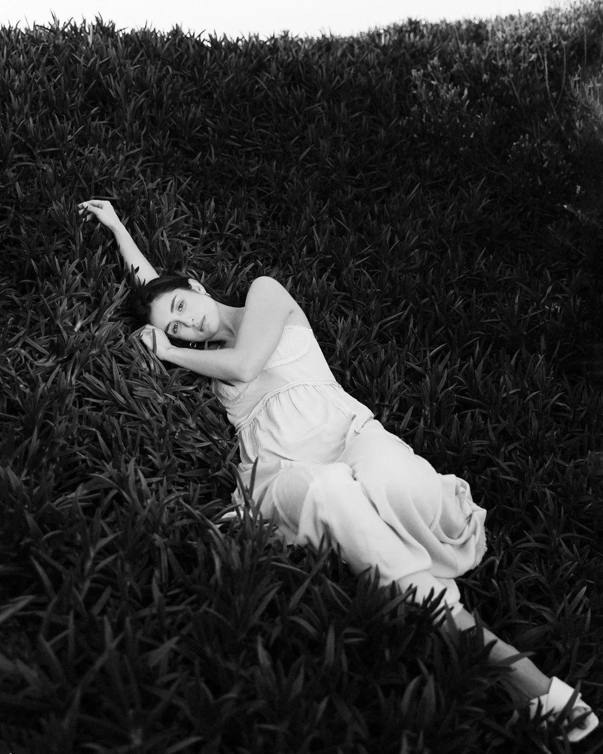 Mulher deitada em meio a folhas, com expressão tranquila, segurando a cabeça com uma mão.
Woman lying among leaves, with a calm expression, holding her head with one hand.