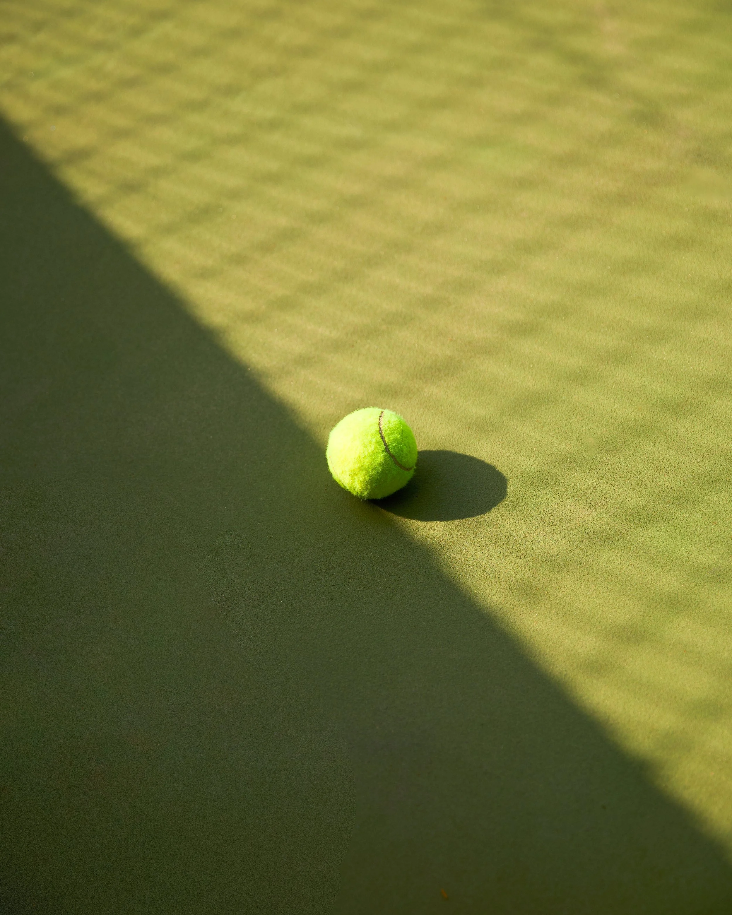 A green tennis ball on a tennis court, with part of the court shaded and part illuminated by sunlight.