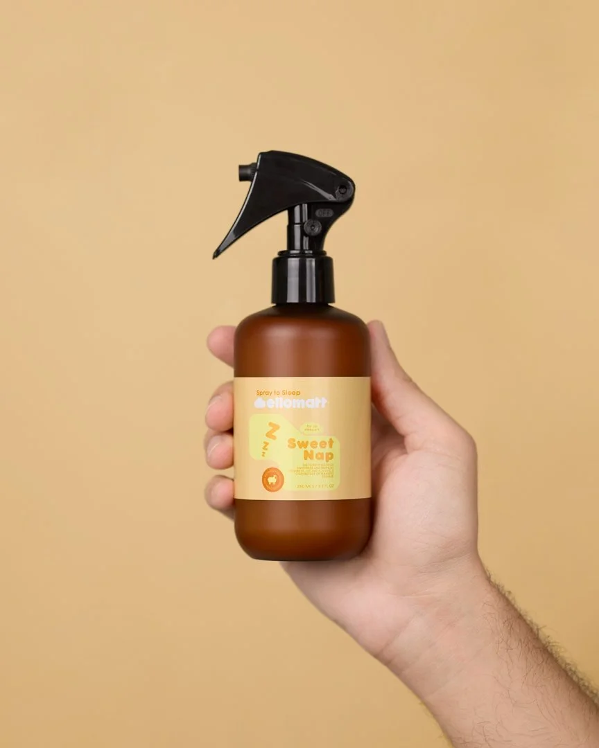 A hand holding a brown spray bottle labeled 'Sweet Nap' with a black spray nozzle, against a beige background.