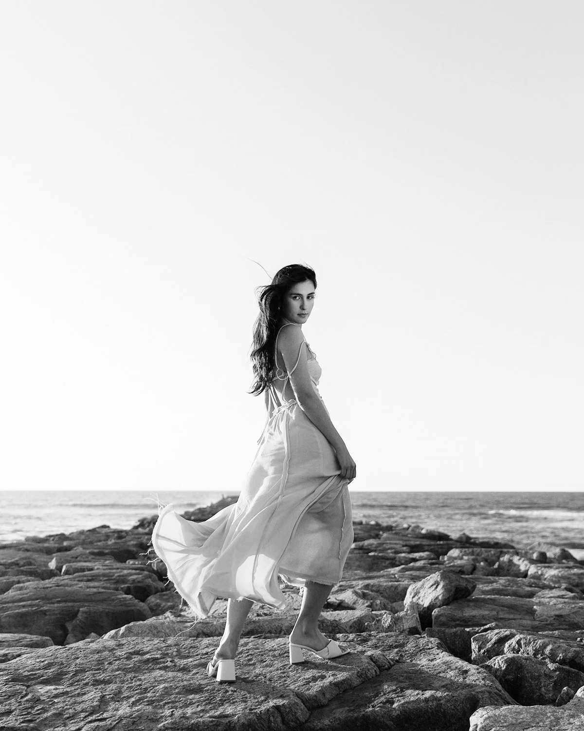 Mulher de vestido fluido em uma praia rochosa, no pôr do sol, em preto e branco.
Woman in flowing dress on a rocky beach at sunset in black and white.