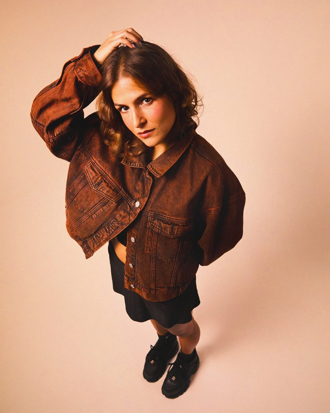 A woman with brown hair wearing a brown denim jacket, black skirt, and black sneakers, posing against a beige background.