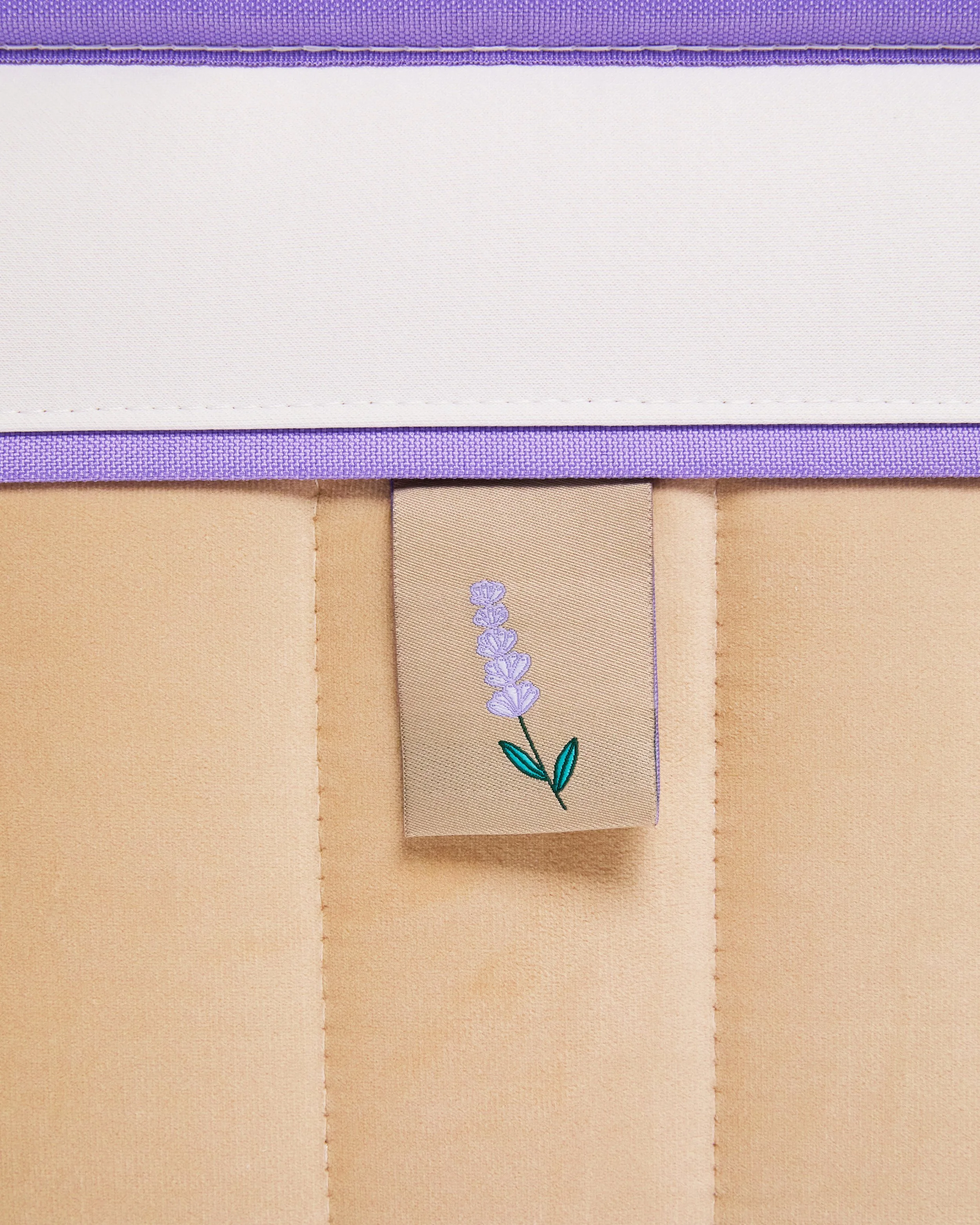 Close-up of a fabric corner with a lavender flower embroidered on a beige tag hanging from a seam, with purple and white fabric borders.