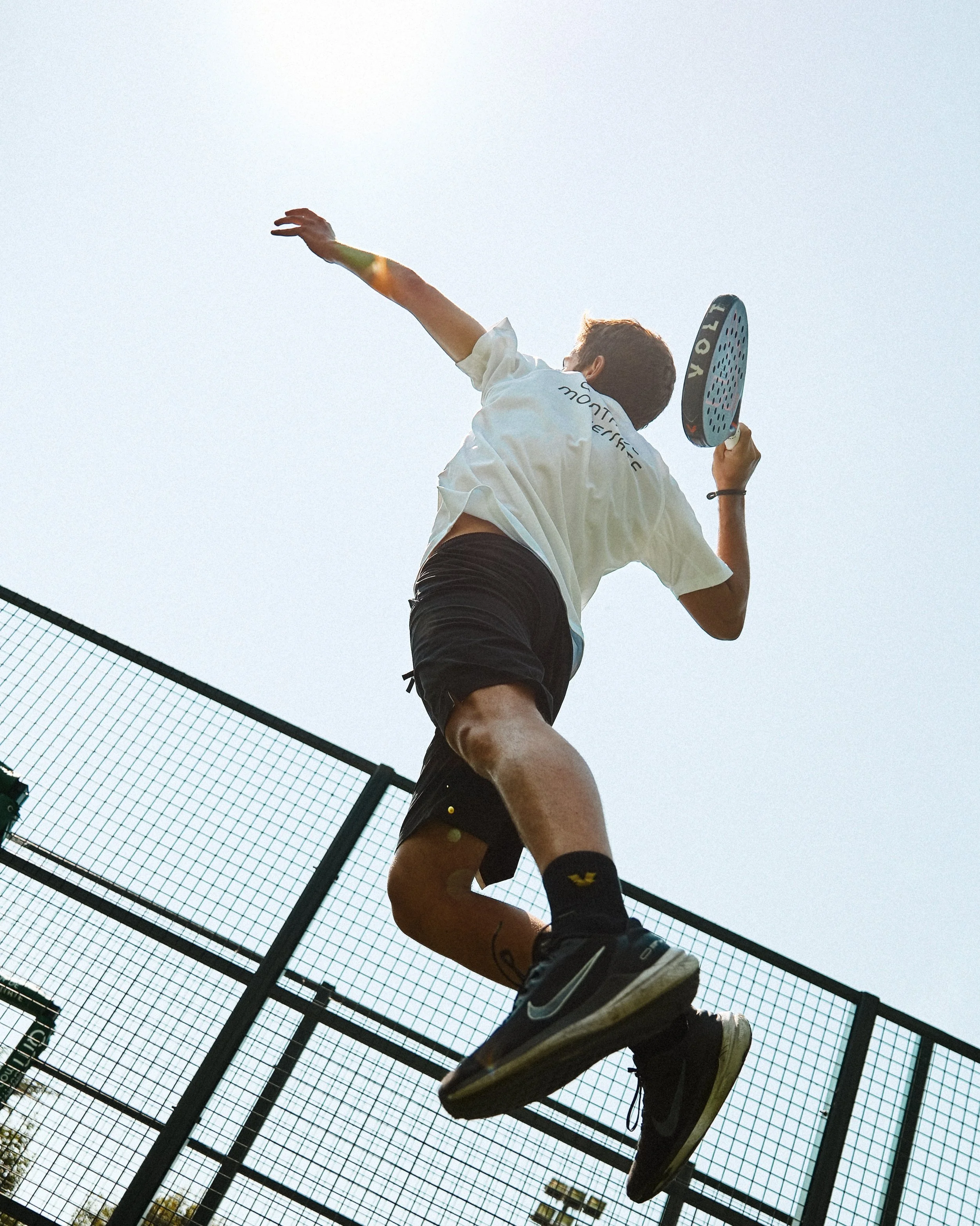 A person playing pickleball on an outdoor court, jumping in the air with a paddle in hand, seen from a low angle against a clear sky.