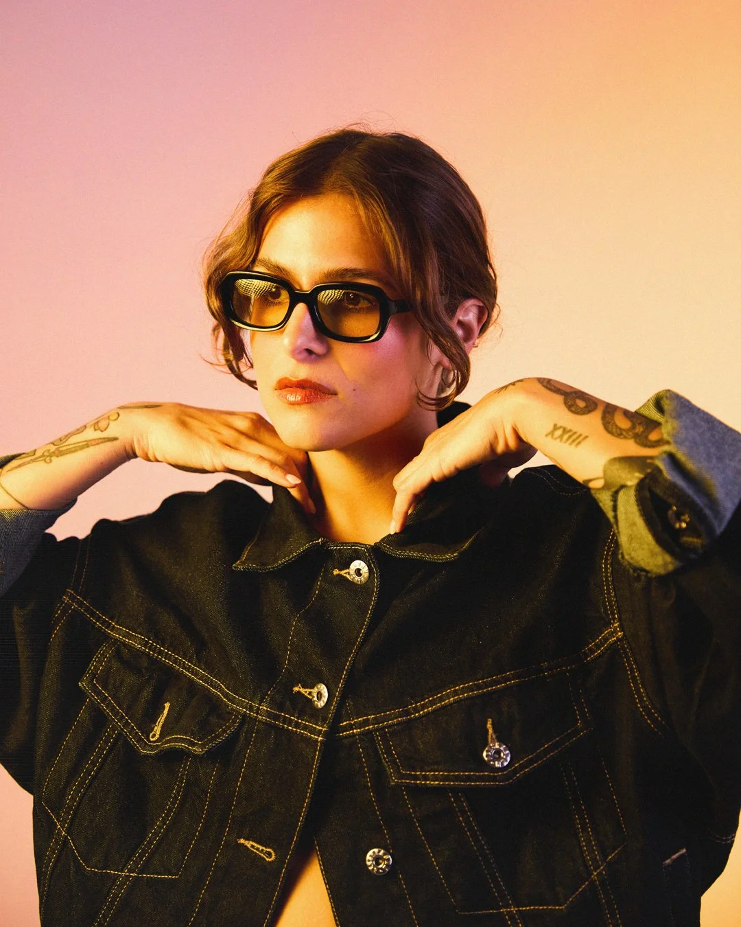 A young woman with short brown hair wearing black sunglasses, a black denim jacket with yellow stitching, and tattoos on her arms, posing with hands near her face against a pastel-colored background.