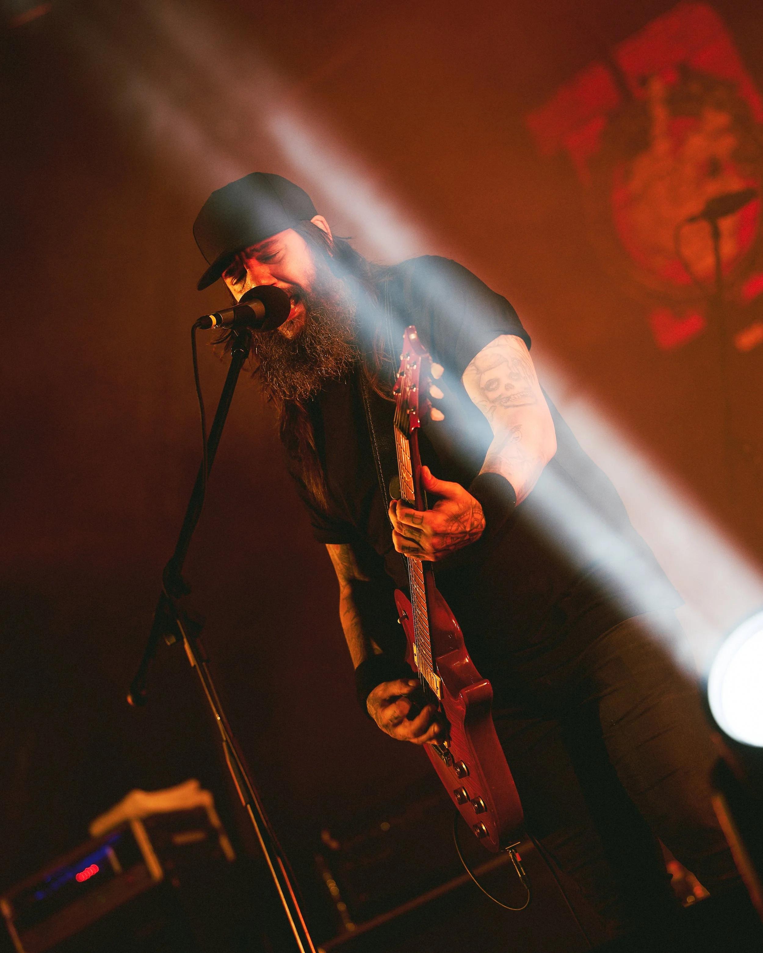Musician performing on stage with red electric guitar, wearing black hat, black t-shirt, and tattoos visible on arms, illuminated by stage lights.