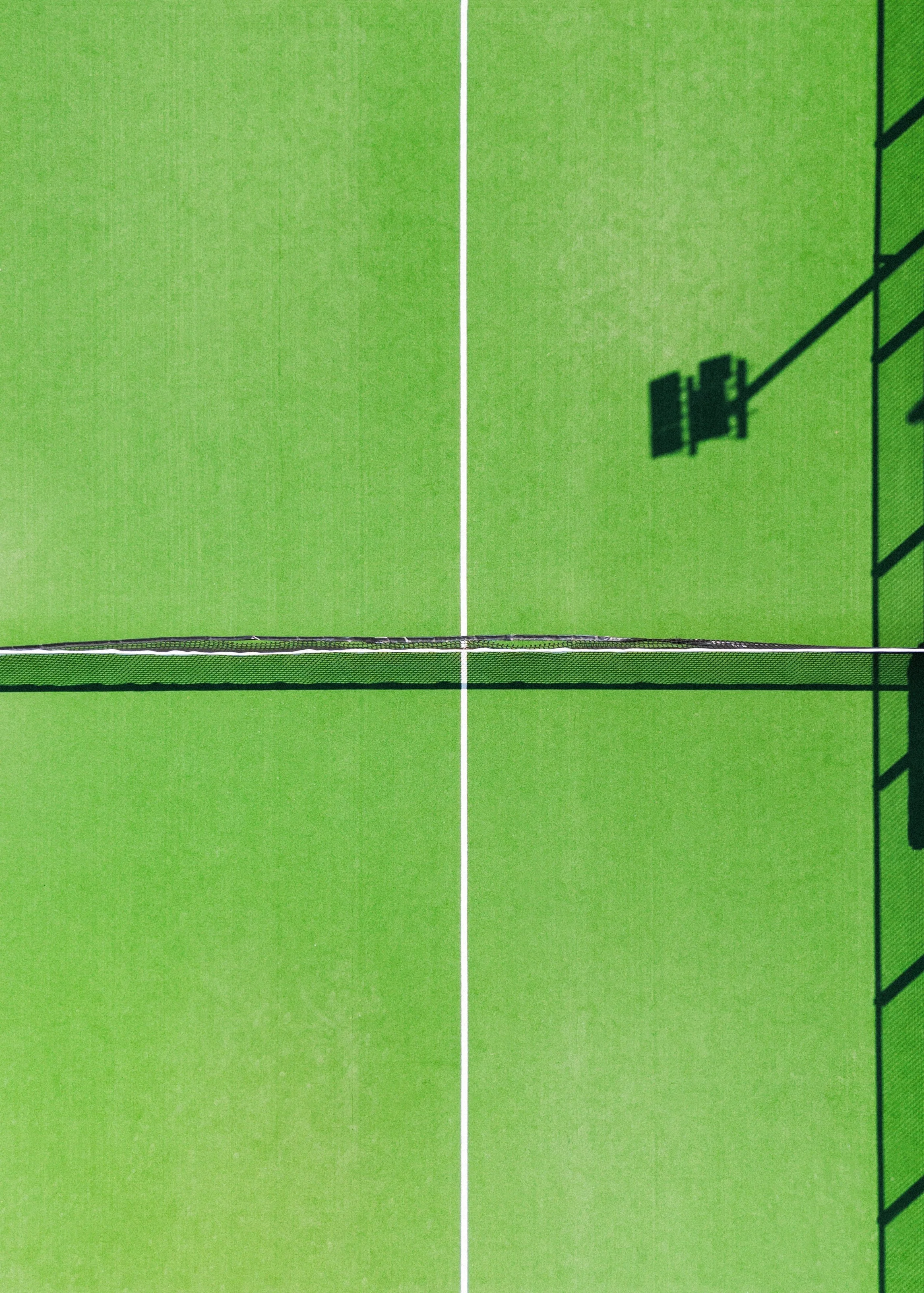 Top-down view of a green tennis court with a black net in the middle, white boundary lines, and a shadow of a tennis court light on the right side.