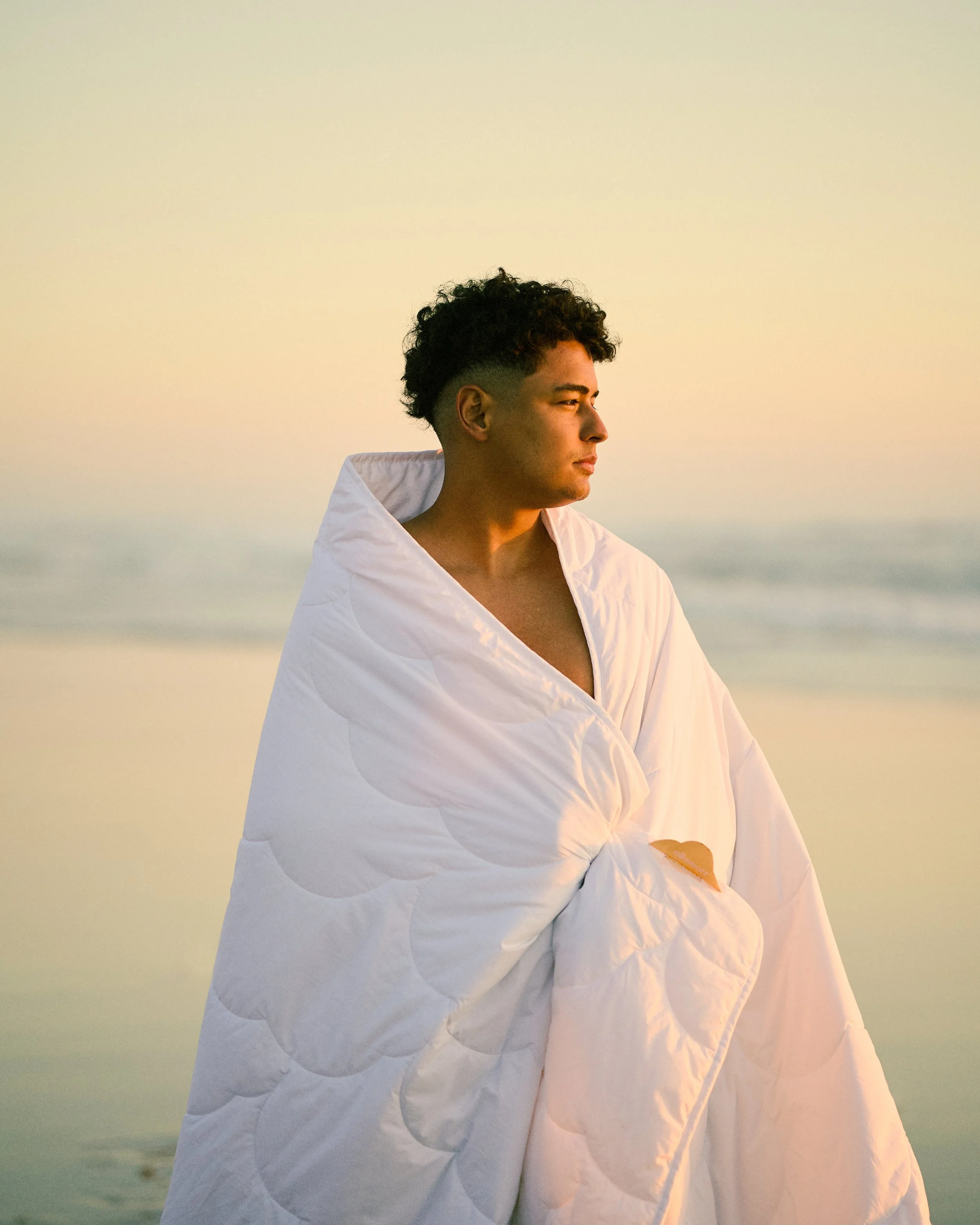 A young man wrapped in a white quilt looking out at the ocean at sunset.