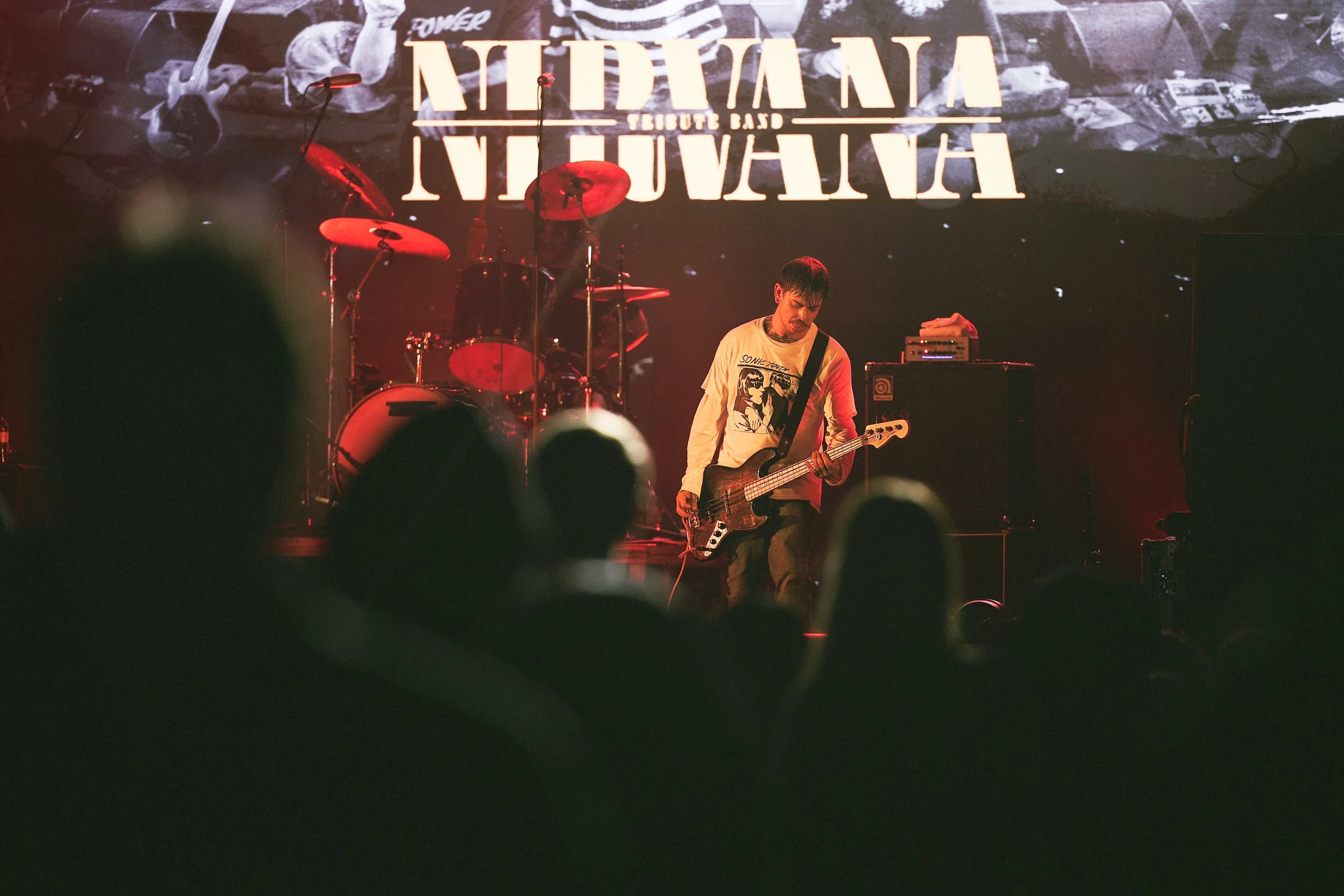 Musician playing bass guitar on stage during a live concert with a drum set and a backdrop displaying text.
