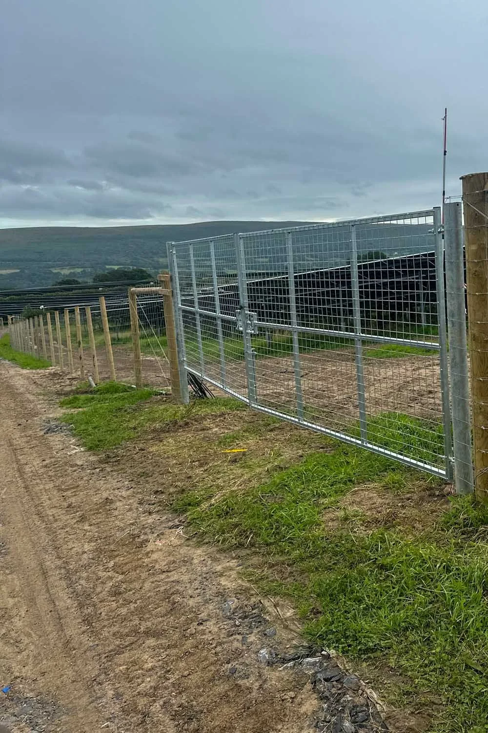 A newly installed metal and wire fence alongside a dirt dirt path, with a cloudy sky and rolling hills in the background.