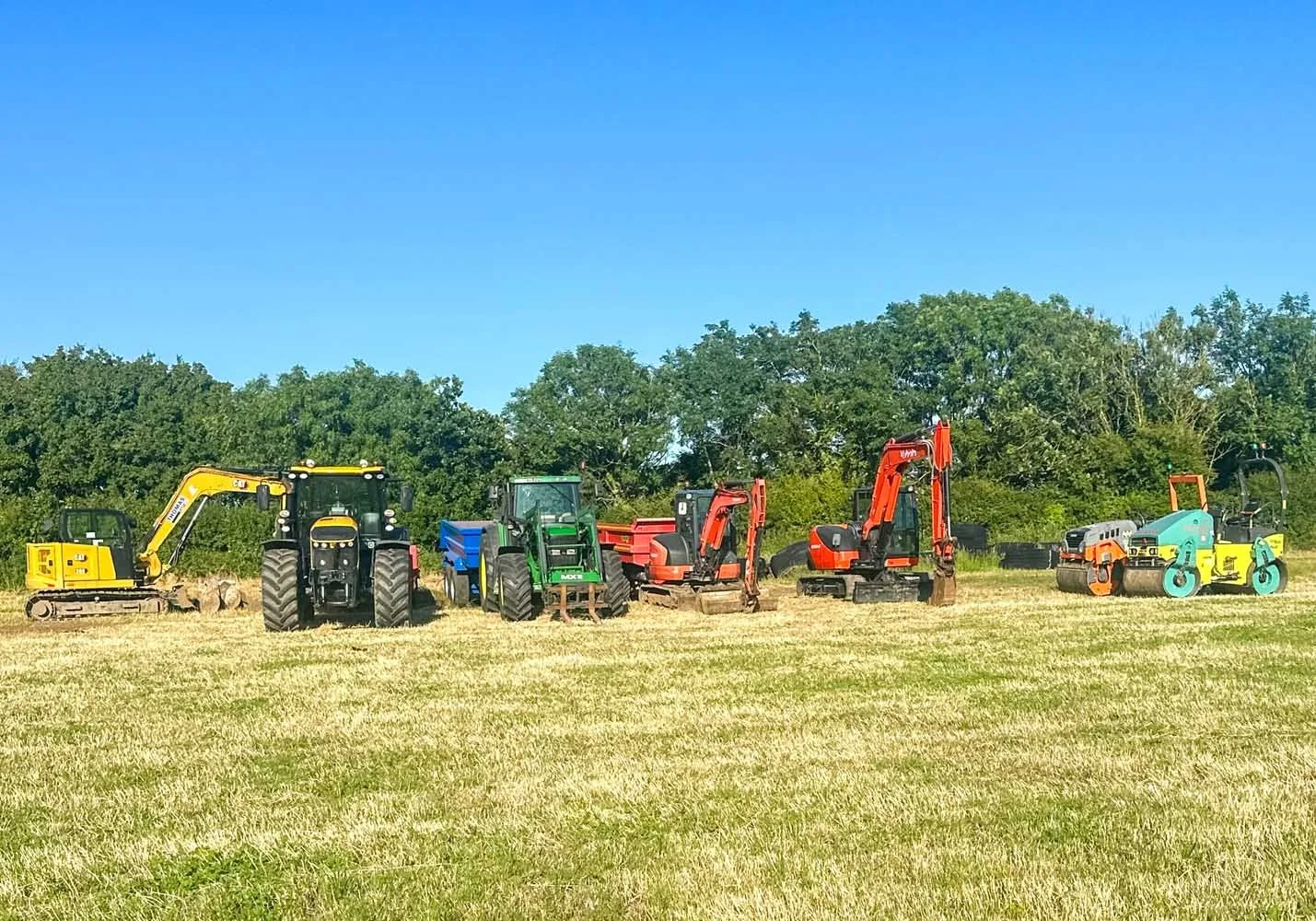 Line of various construction and farming equipment including mini excavators, bulldozers, and a tractor on a grassy field with trees in the background under a clear blue sky.