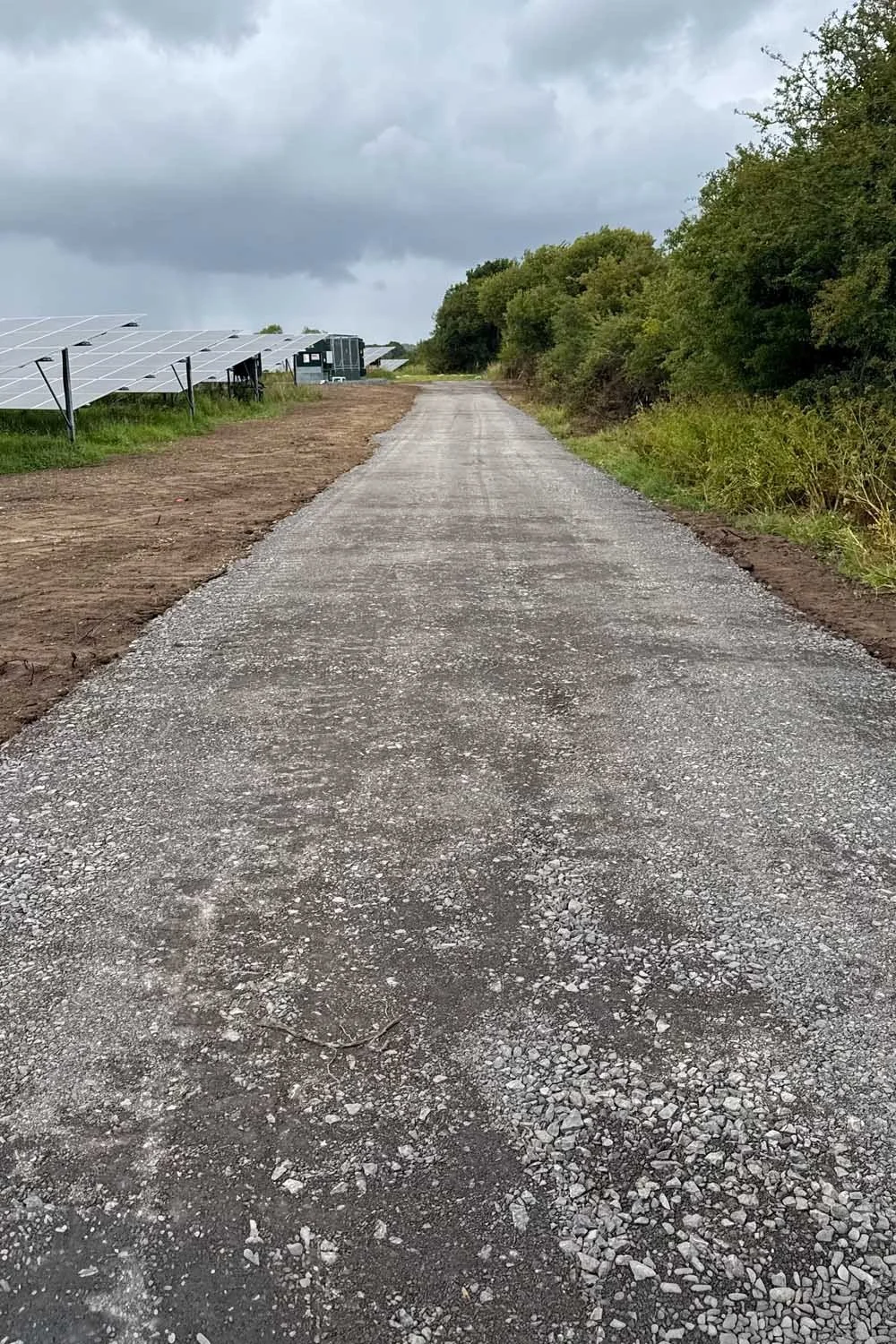 A gravel road running through a rural area with solar panels on the left and trees on the right, under a cloudy sky.