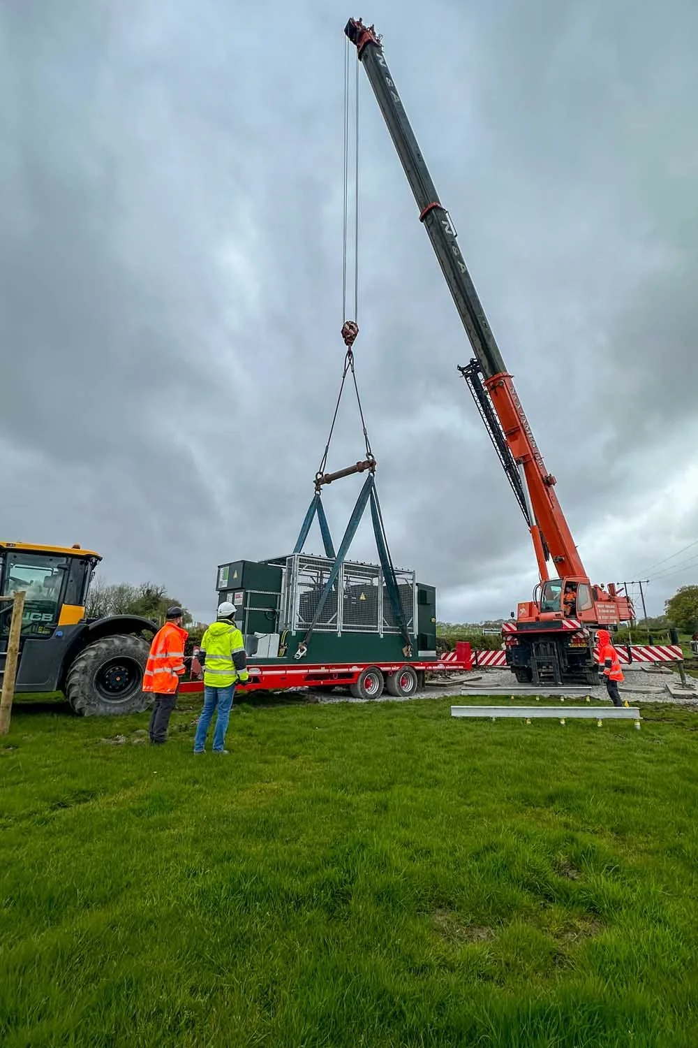 Crane lifting a large industrial generator onto a trailer with four workers observing, one in a yellow vest, two in orange vests, and a person in a red jacket nearby, under a cloudy sky.