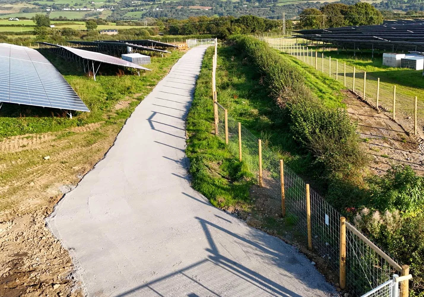 A newly paved concrete pathway through a rural area with solar panel installations on the left and green fields with bushes and trees on the right, surrounded by a wooden and wire fence.