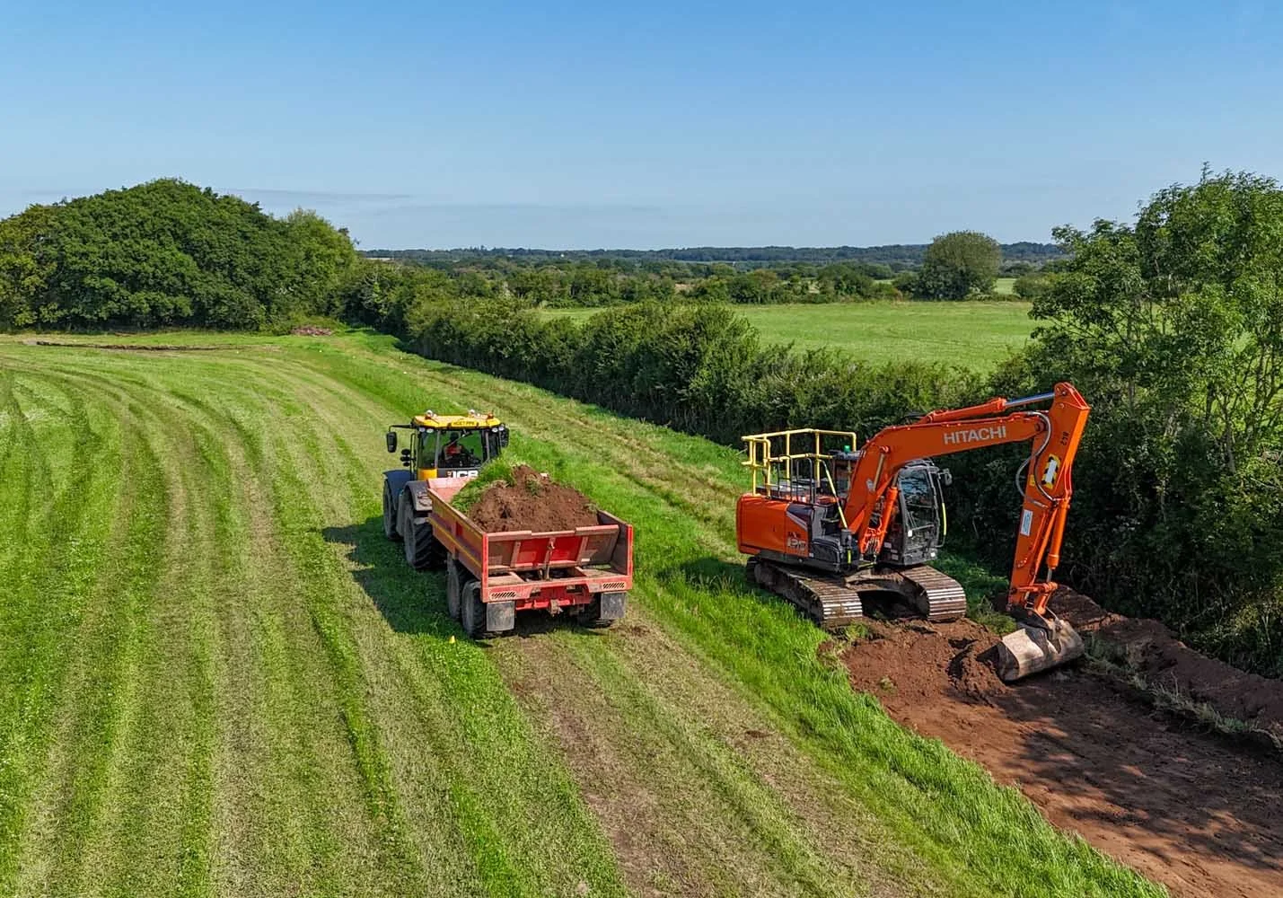 An aerial view of a construction site on a grassy field with a small orange excavator digging a trench and a yellow dump truck transporting soil. The field has green grass with trees and a blue sky in the background.