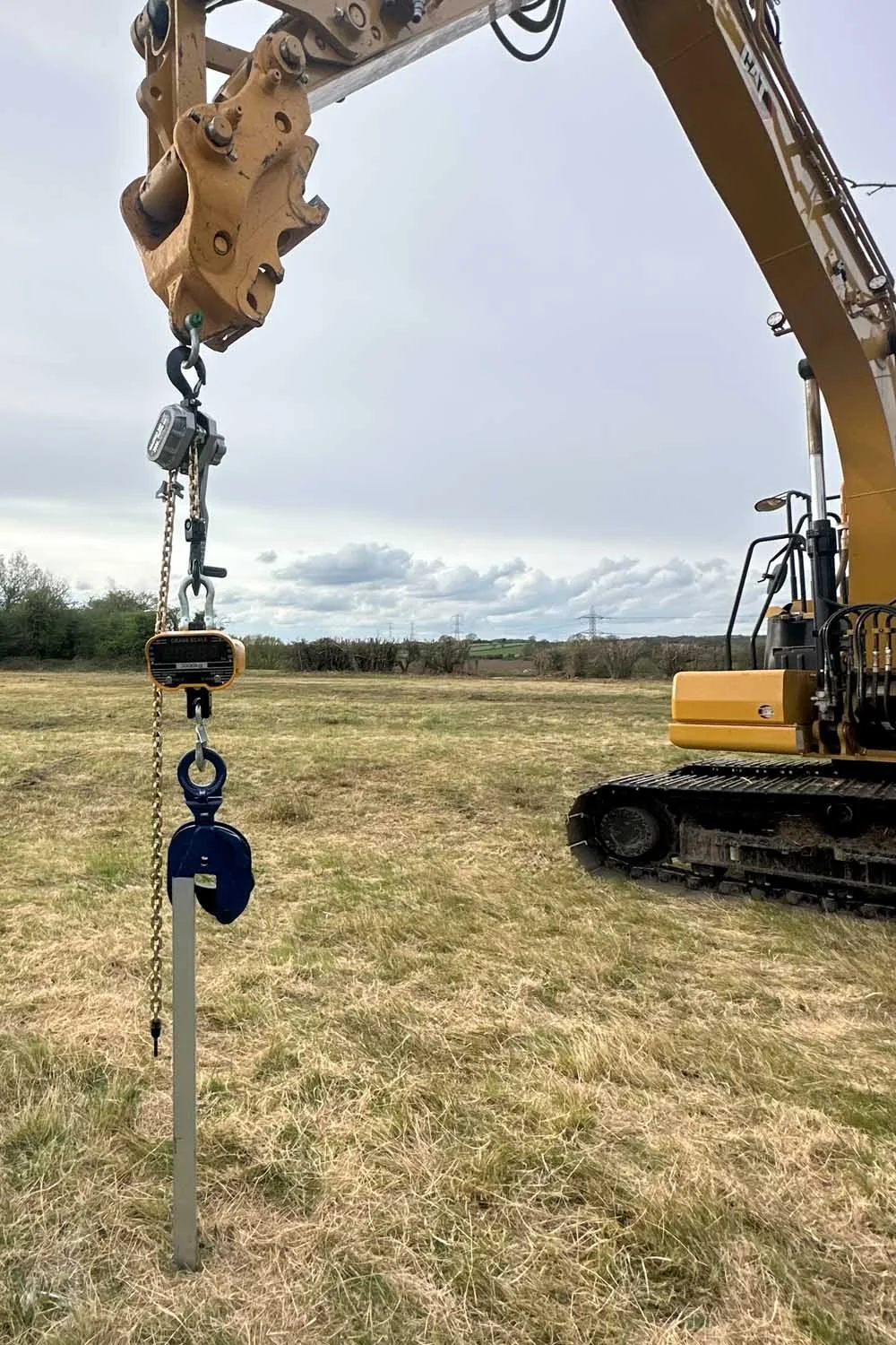 A large yellow excavator with a tracked wheel base, operating in a field with a sketchy sky. The excavator’s arm extends over a metal marker pole with construction tools hanging on it, including a chain, hook, level, and other equipment.