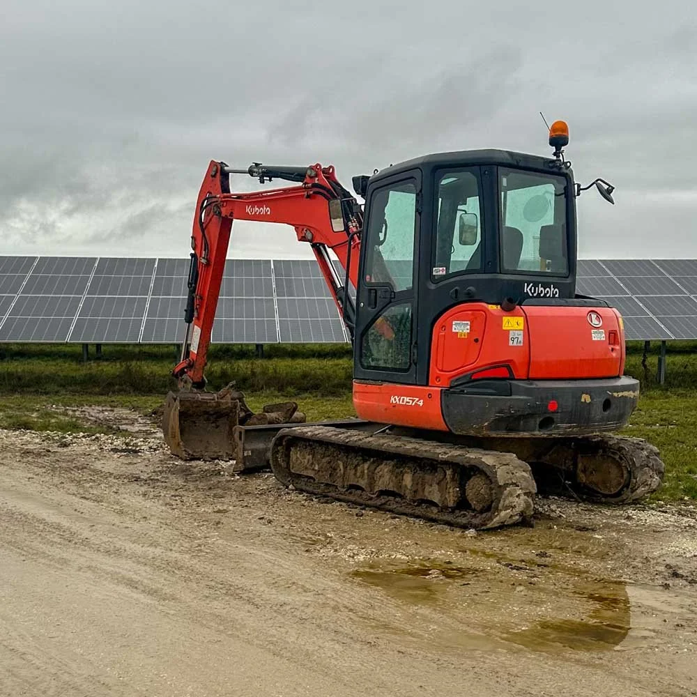 A small Kubota excavator sits on a dirt road with a solar panel array in the background and a cloudy sky overhead.