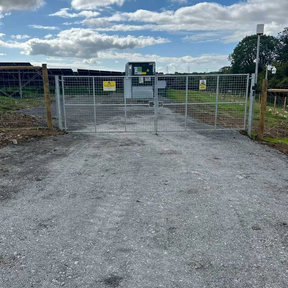 Gravel driveway leading to a fenced electric security gate at a solar farm with solar panels, warning signs, and a gray control box under a partly cloudy sky.