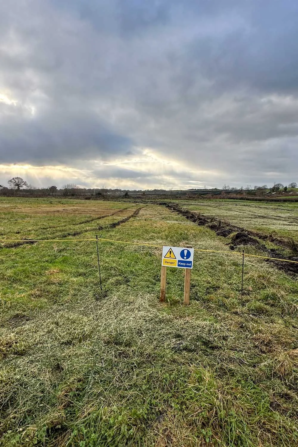 A grassy field with dirt tracks and a warning sign that says 'Danger' and 'Keep out,' surrounded by a thin yellow rope under a cloudy sky.