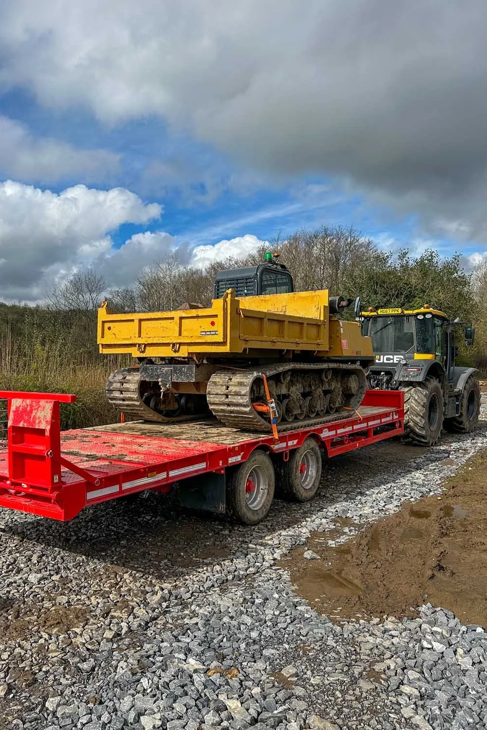 A yellow tracked utility vehicle loaded onto a red flatbed trailer at a construction site with gravel and dirt, and a background of trees and a cloudy sky.