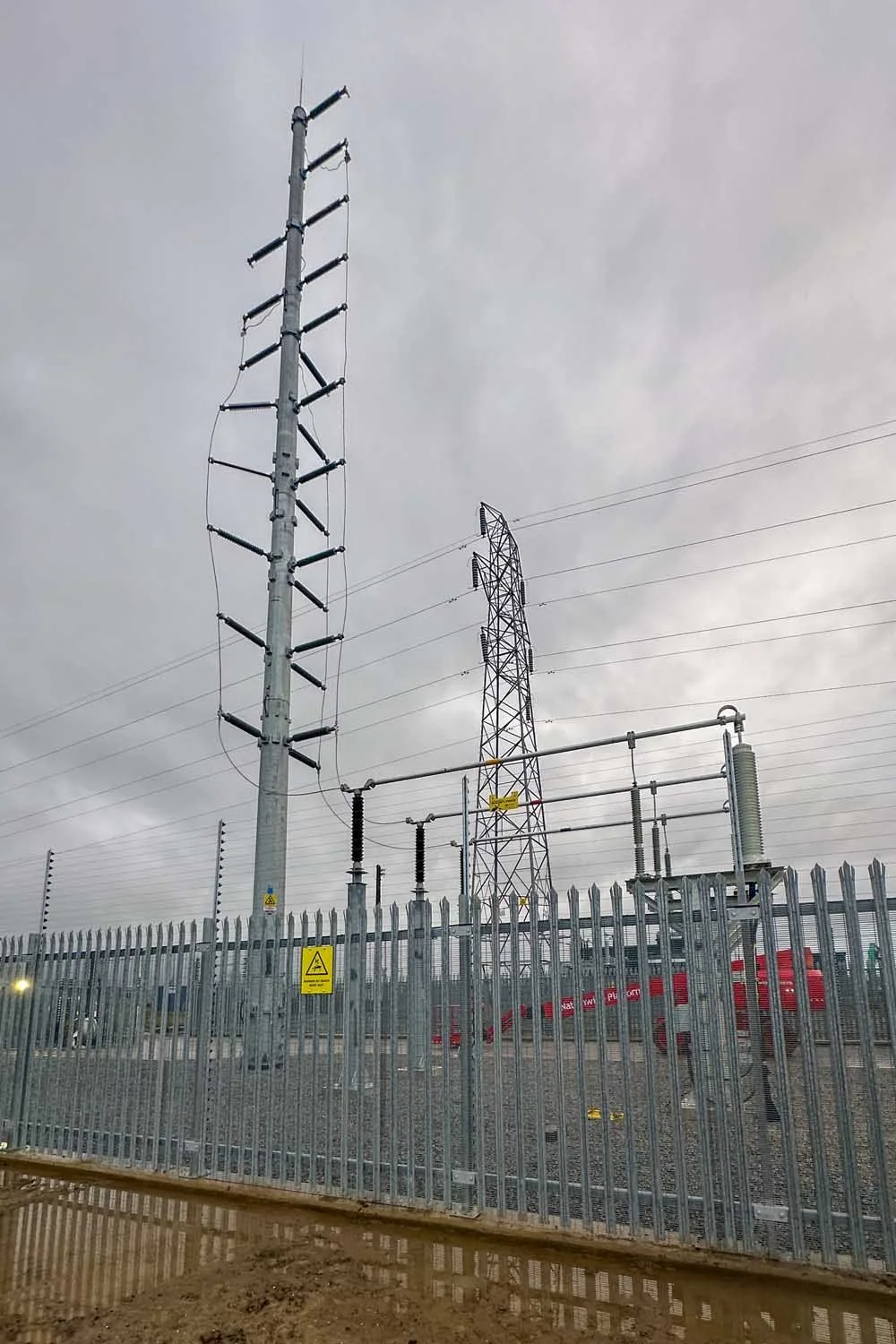 Electricity transmission towers and equipment behind a metal fence under cloudy sky.