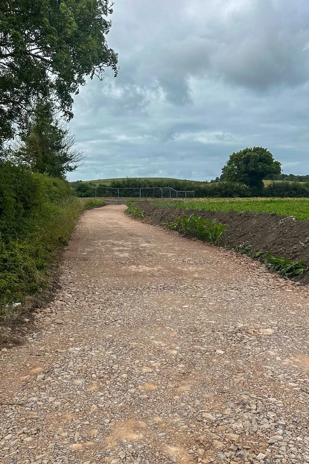 A gravel country road winding through green fields and trees under a cloudy sky.