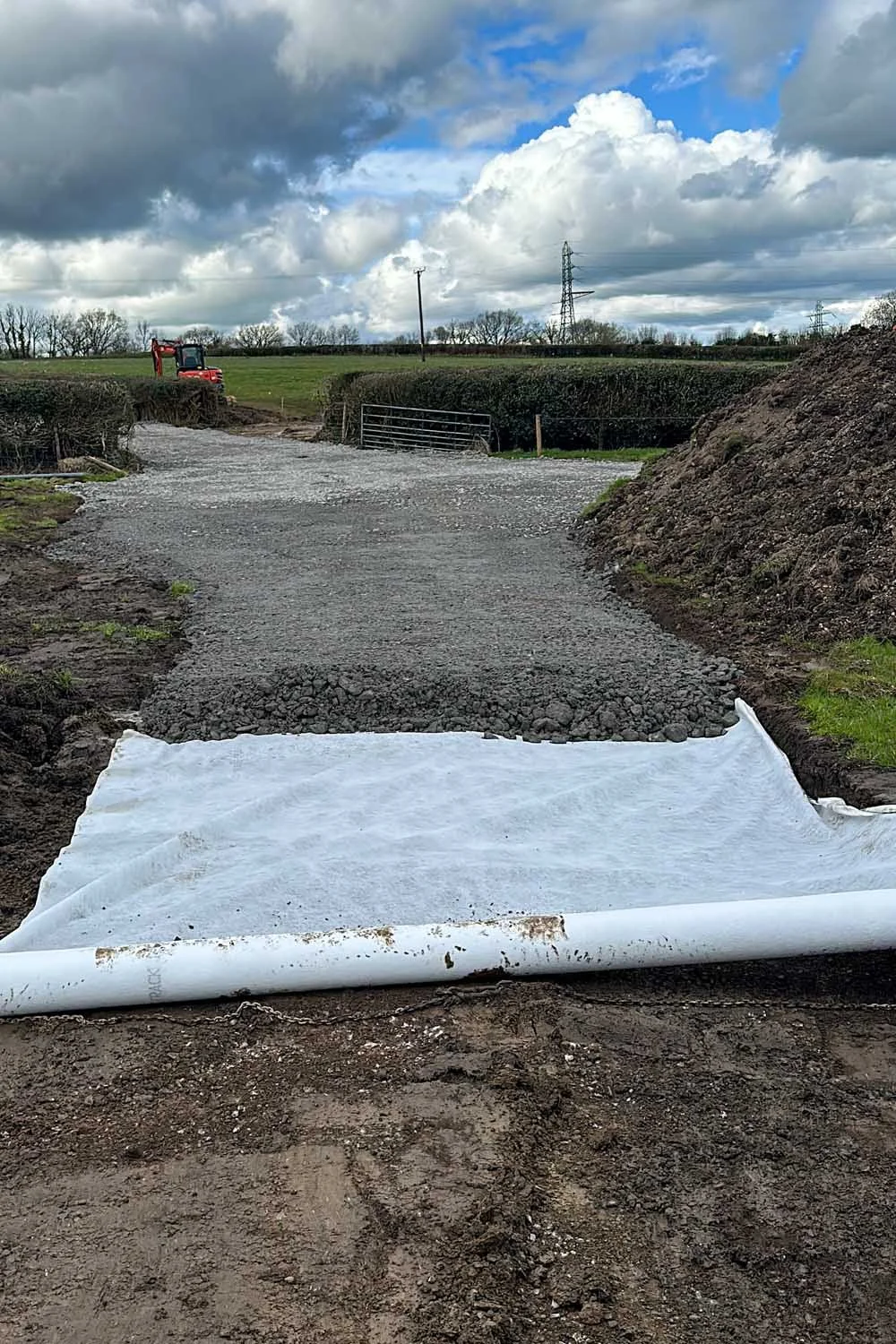A rural dirt and gravel road under construction with a white geotextile fabric laid on the ground, surrounded by green fields, hedges, and a red tractor in the distance, with a cloudy sky overhead.