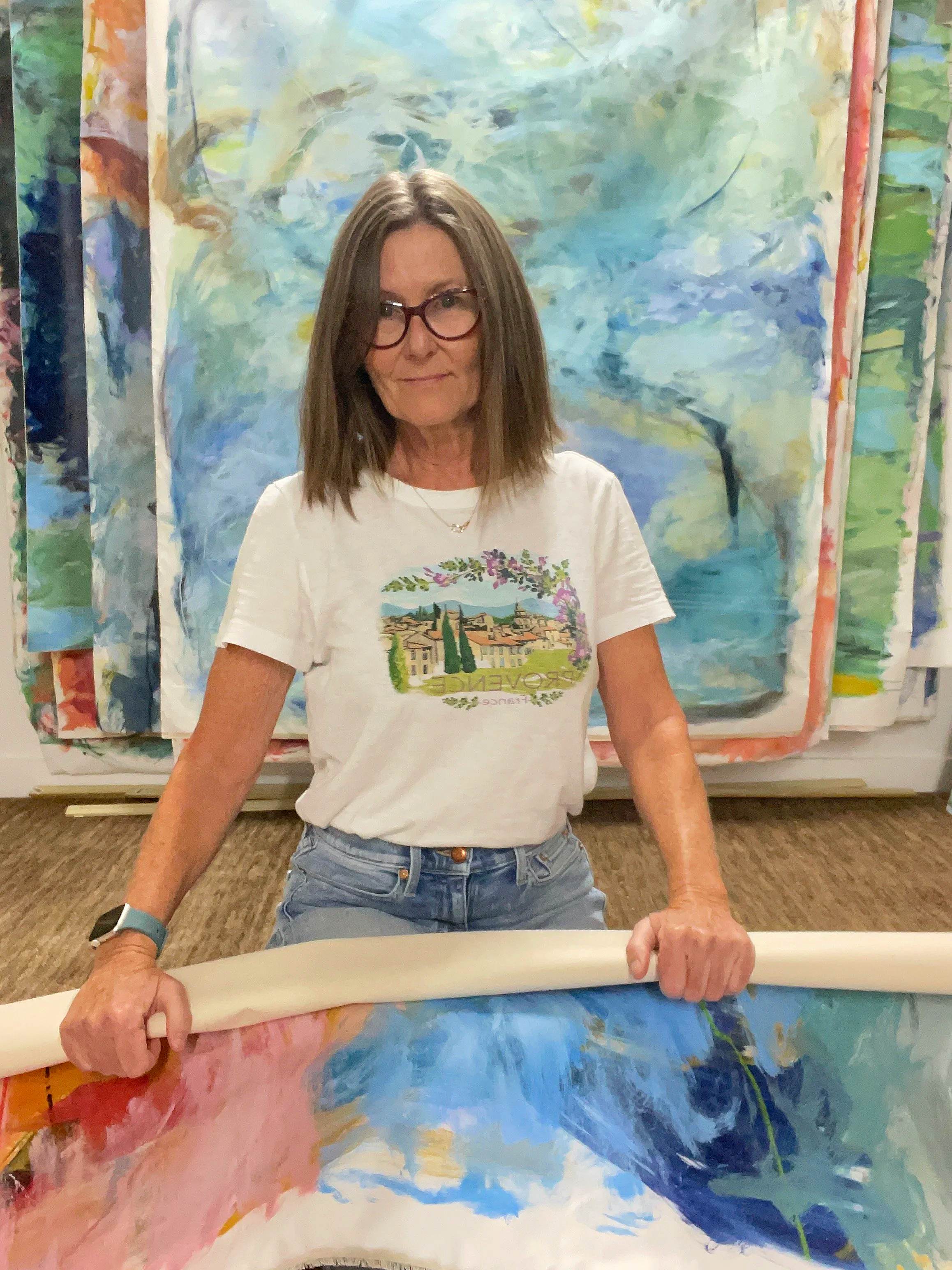 A woman with brown hair, glasses, and a white T-shirt featuring a landscape design, holding a long white roller brush in front of a large abstract painting with blue, green, and pink brushstrokes.