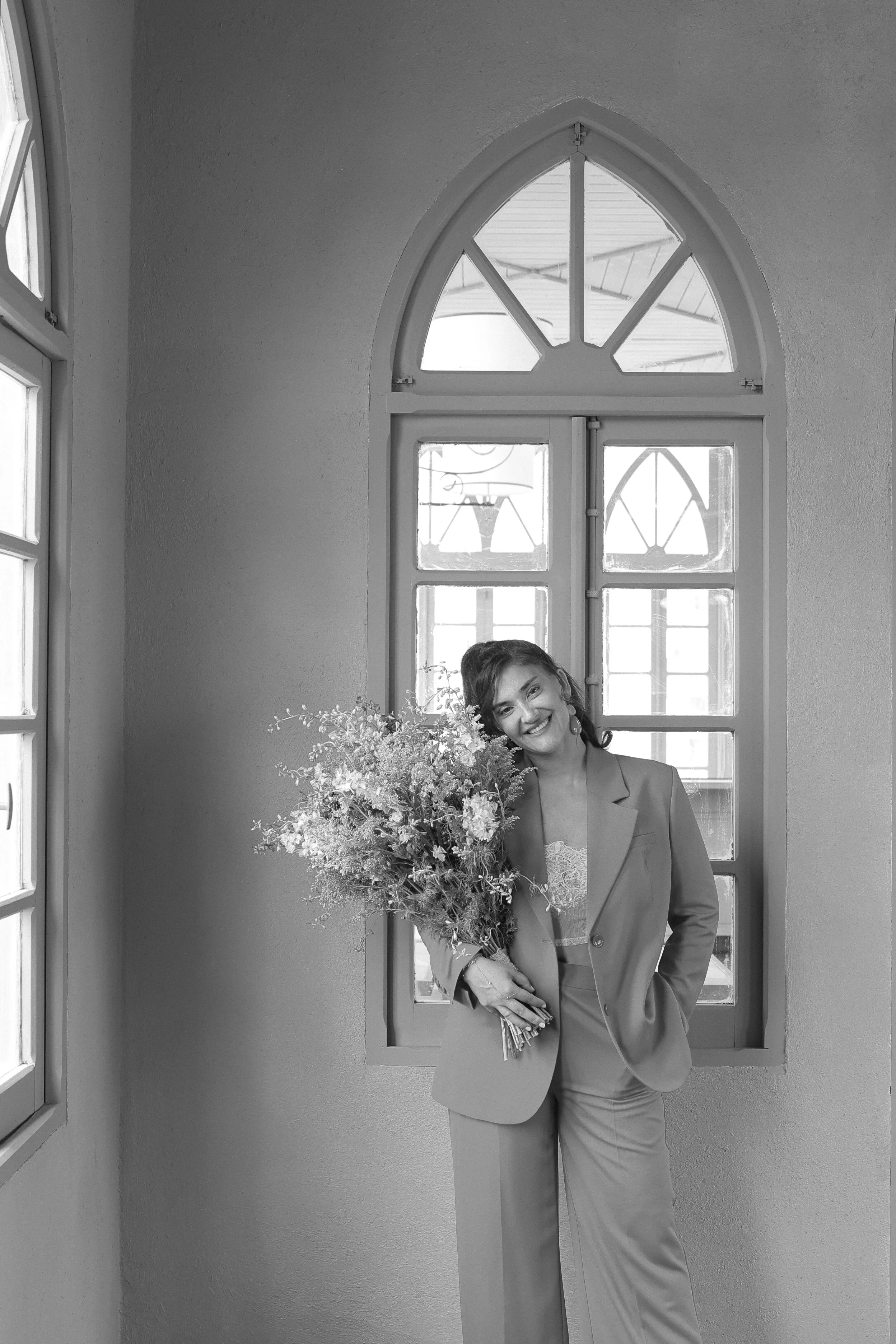 A woman in a suit holding a large bouquet of flowers, smiling, standing in front of a pointed arch window in a room with a textured wall.